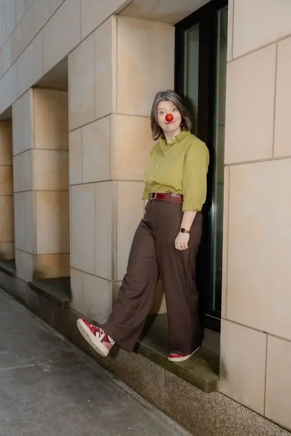 Frau steht auf einem Sims vor einer Wand, trägt eine rote Clownsnase und rote Sneakers, die wie Sneakers aussehen.