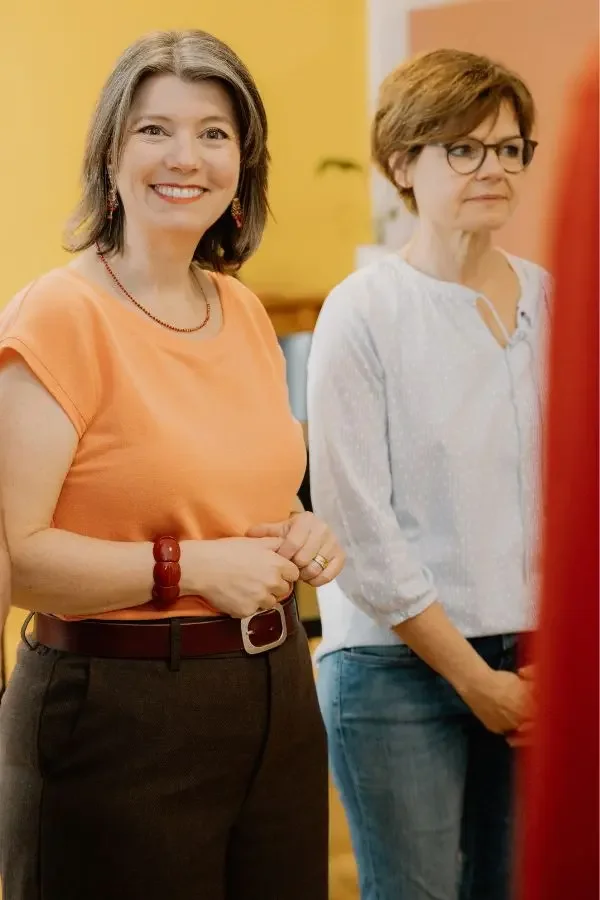 Zwei Frauen stehen nebeneinander in einem Raum mit gelbem Hintergrund, eine lächelnd, die andere nachdenklich.