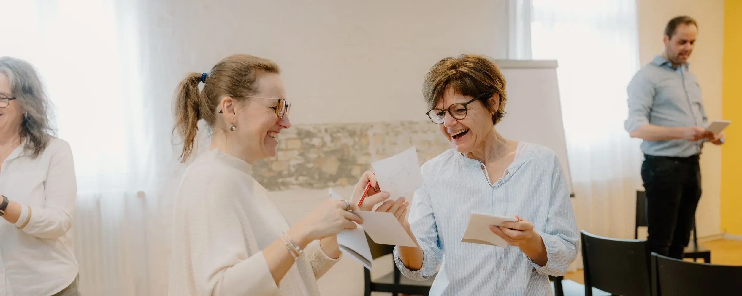 Drei Frauen lachen und unterhalten sich beim Lesen von Briefen in einem hellen Raum, im Hintergrund ein Mann, der ein Papier hält.
