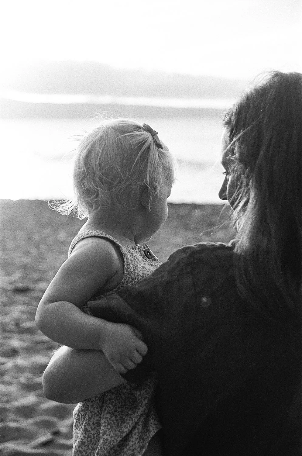 A child and an adult with light and dark hair, respectively, holding each other at the beach, facing the ocean during sunset in black and white.