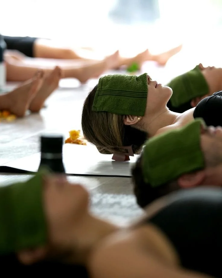 People lying on their backs during a relaxation or meditation session, with green towels over their eyes in a calm environment.