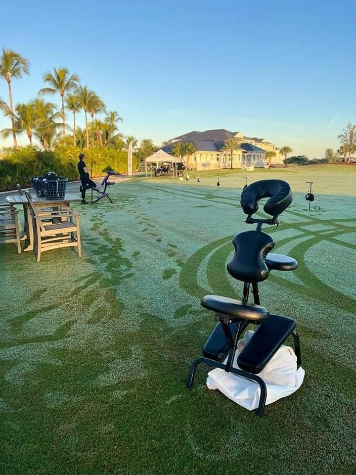 A golf course with a massage chair in the foreground, a person exercising on a stationary bike, and a large house in the background, surrounded by palm trees under a clear sky.