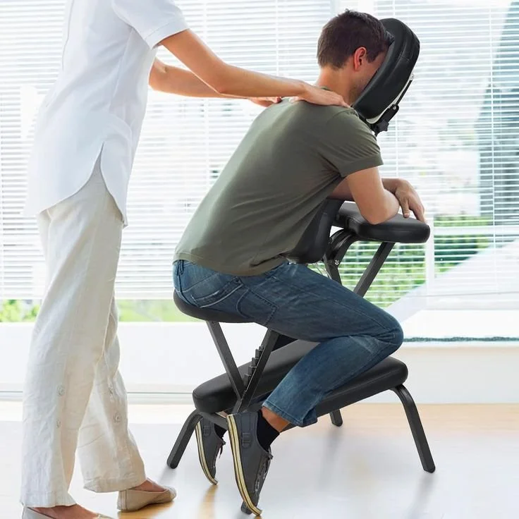 A person receiving a chair massage from a massage therapist in a room with large windows and horizontal blinds.