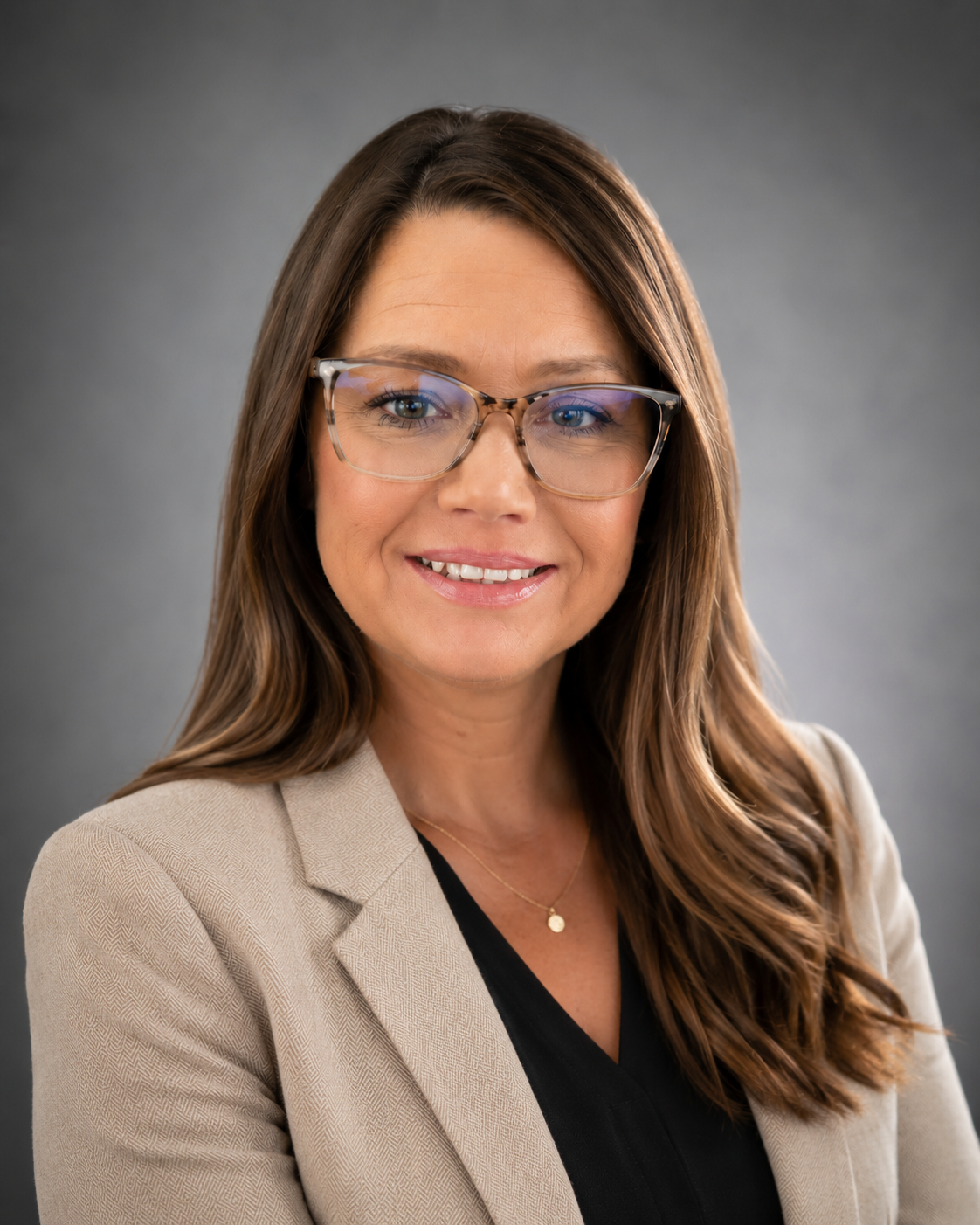 A woman with long brown hair, wearing glasses, a beige blazer, and a black top, smiling at the camera against a gray background.