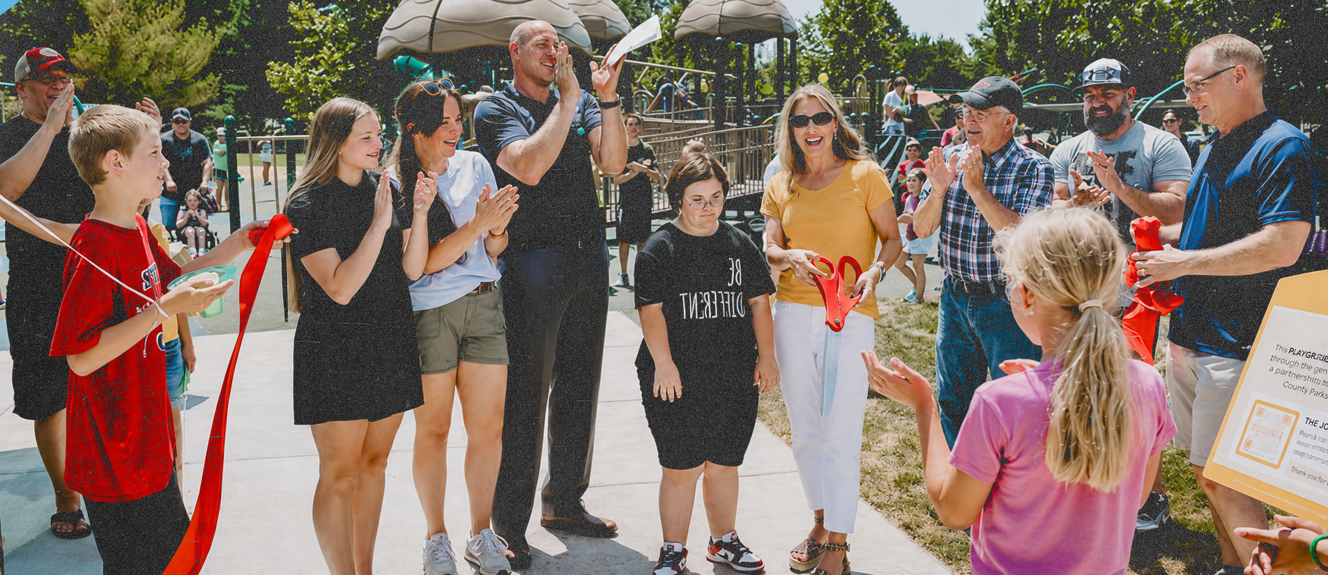 People gathered outdoors at a park celebrating a ribbon-cutting event, with some holding scissors and ribbons, surrounded by children and adults clapping and smiling.