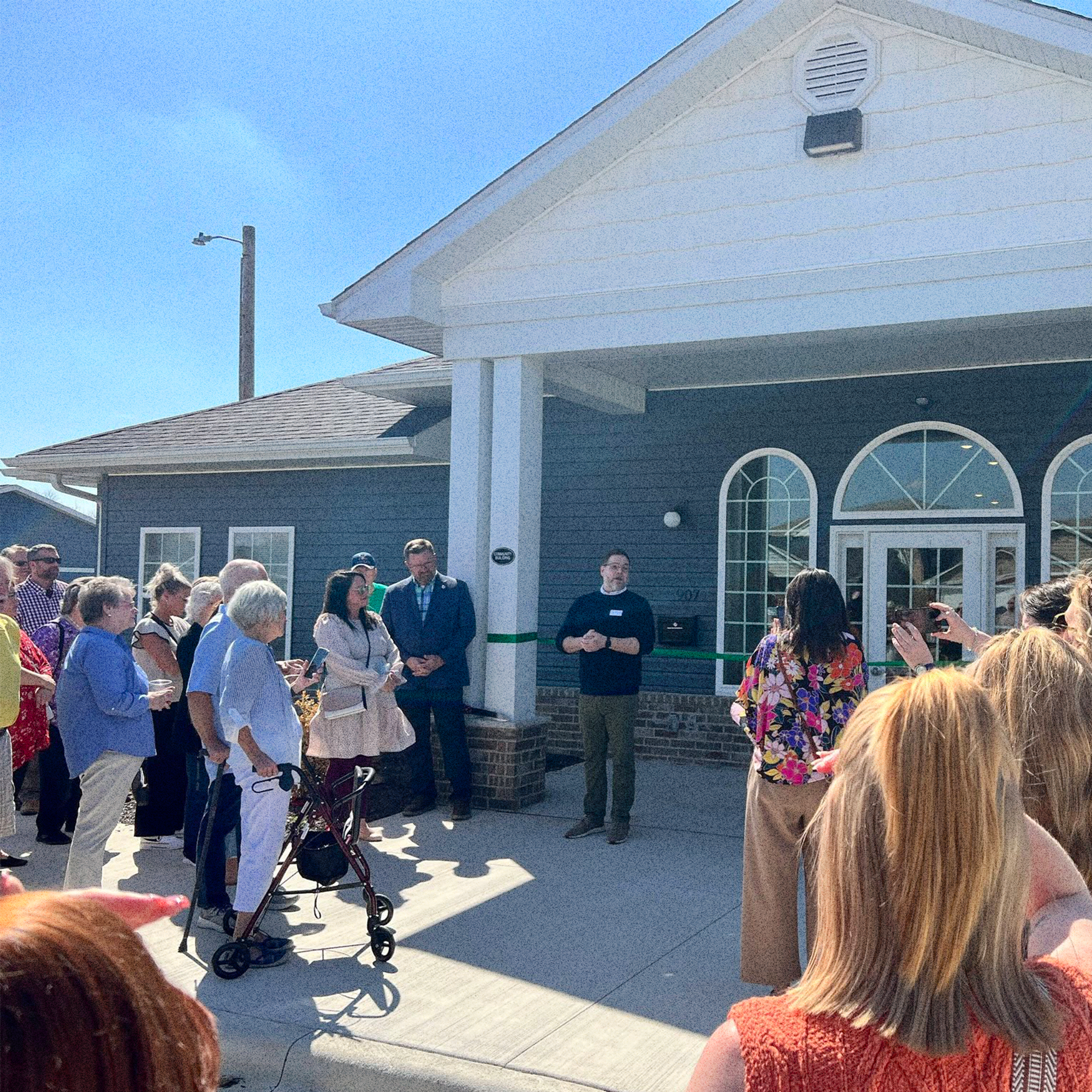 Community gathering at the Generations Village ribbon cutting held outdoors in front of a light blue building on a sunny day.