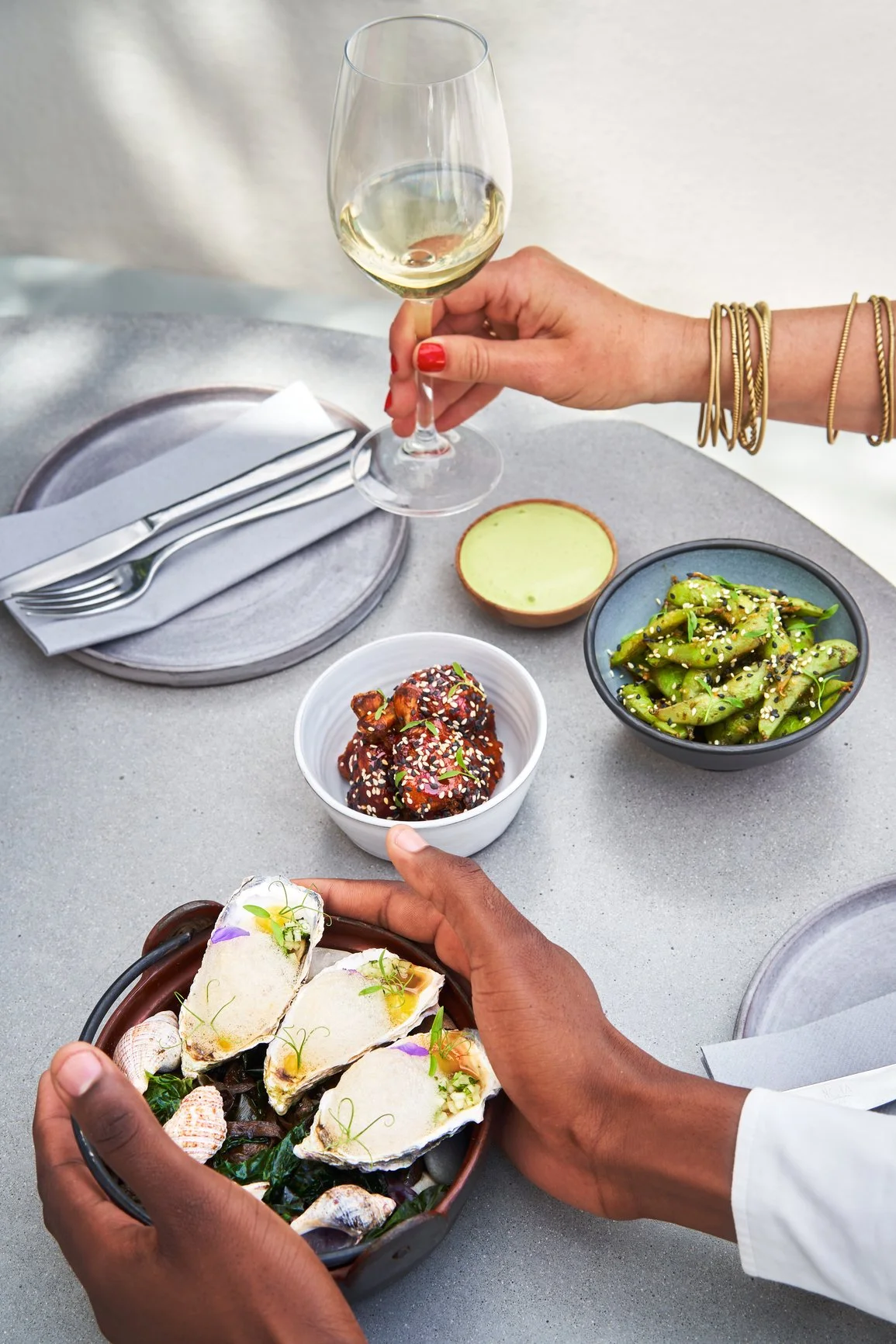 Ridge Haven - New Townhomes for Rent in Concord | A person holding a bowl of oysters, with various dishes and drinks on a table, including a glass of white wine, a bowl of cooked edamame