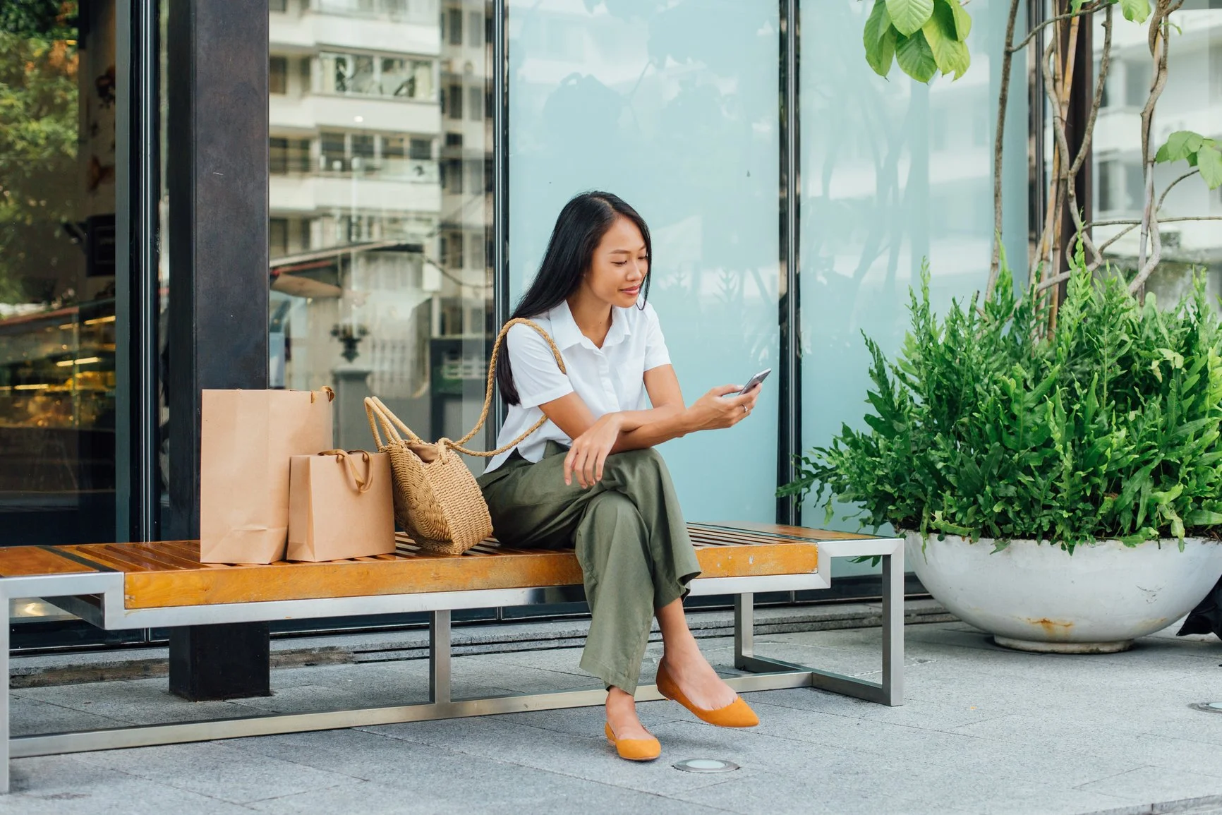 Ridge Haven - New Townhomes for Rent in Concord | A woman sitting on a wooden bench outside a building, looking at her phone, with shopping bags and a woven handbag beside her, and a large potted plant nearby.