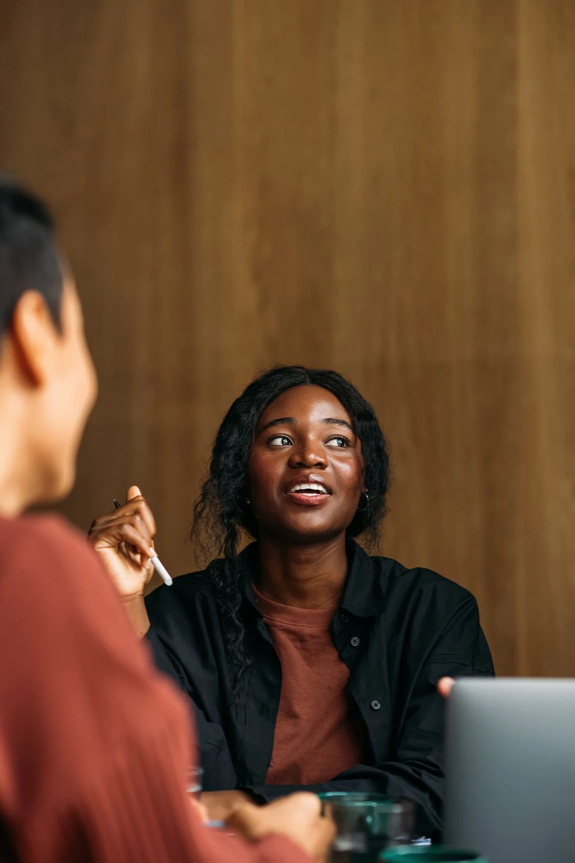 Ridge Haven - New Townhomes for Rent in Concord | A woman with dark curly hair and a black shirt sitting at a table, holding a pen, engaged in conversation with another person whose face and body are partially visible.