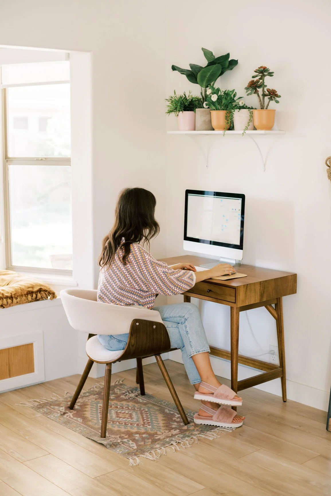 Ridge Haven - New Townhomes for Rent in Concord | A woman working on a desktop computer at a wooden desk in a bright room with a window and potted plants on a wall shelf.