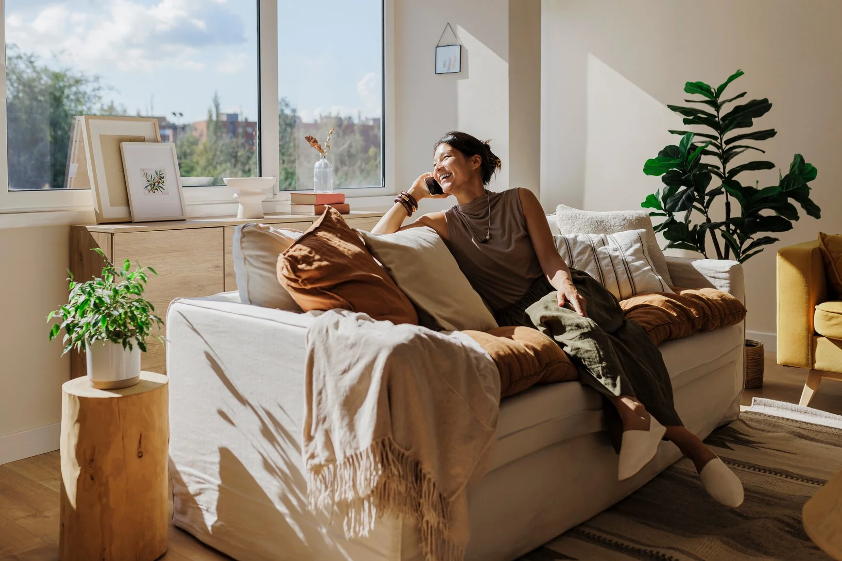 Ridge Haven - New Townhomes for Rent in Concord | A woman sitting on a beige sofa, talking on her cellphone, smiling, in a bright living room with plants, pillows, artwork, and large windows showing an outdoor view.