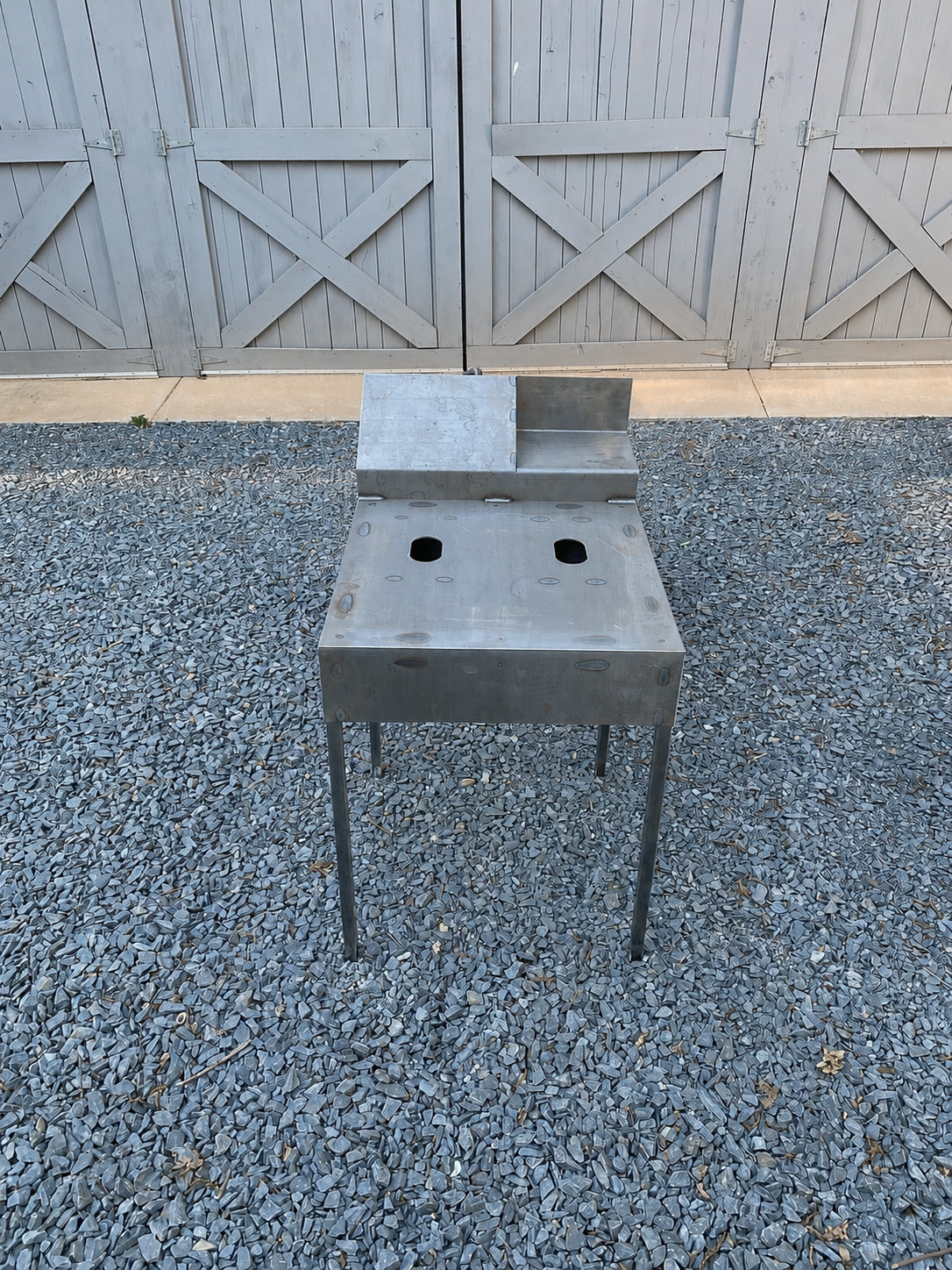 Empty stainless steel outdoor grill on gravel ground with a gray wooden fence in the background.