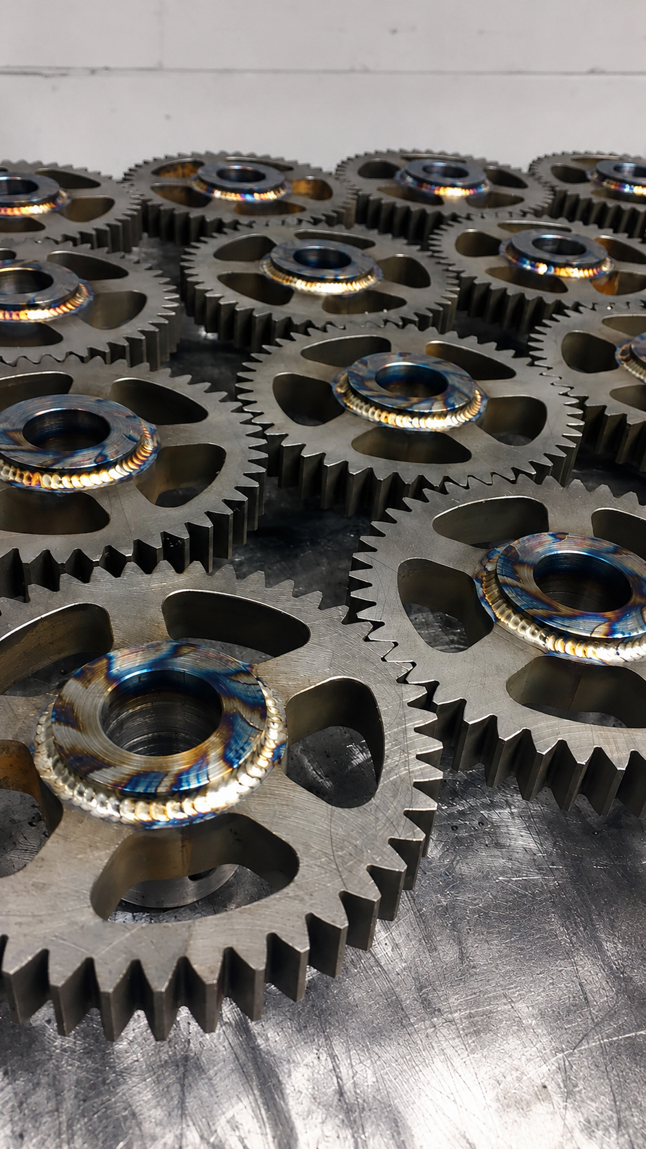 Multiple large metal gears with beveled edges and central holes, lying on a metallic work surface.