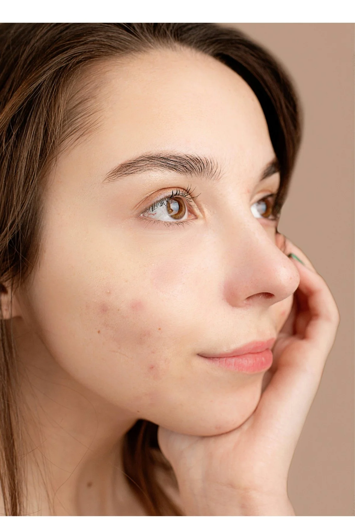 Primer plano de rostro de una joven con piel clara, ojos marrones, cejas definidas y cabello castaño, contemplando hacia la izquierda con mano en mejilla.
