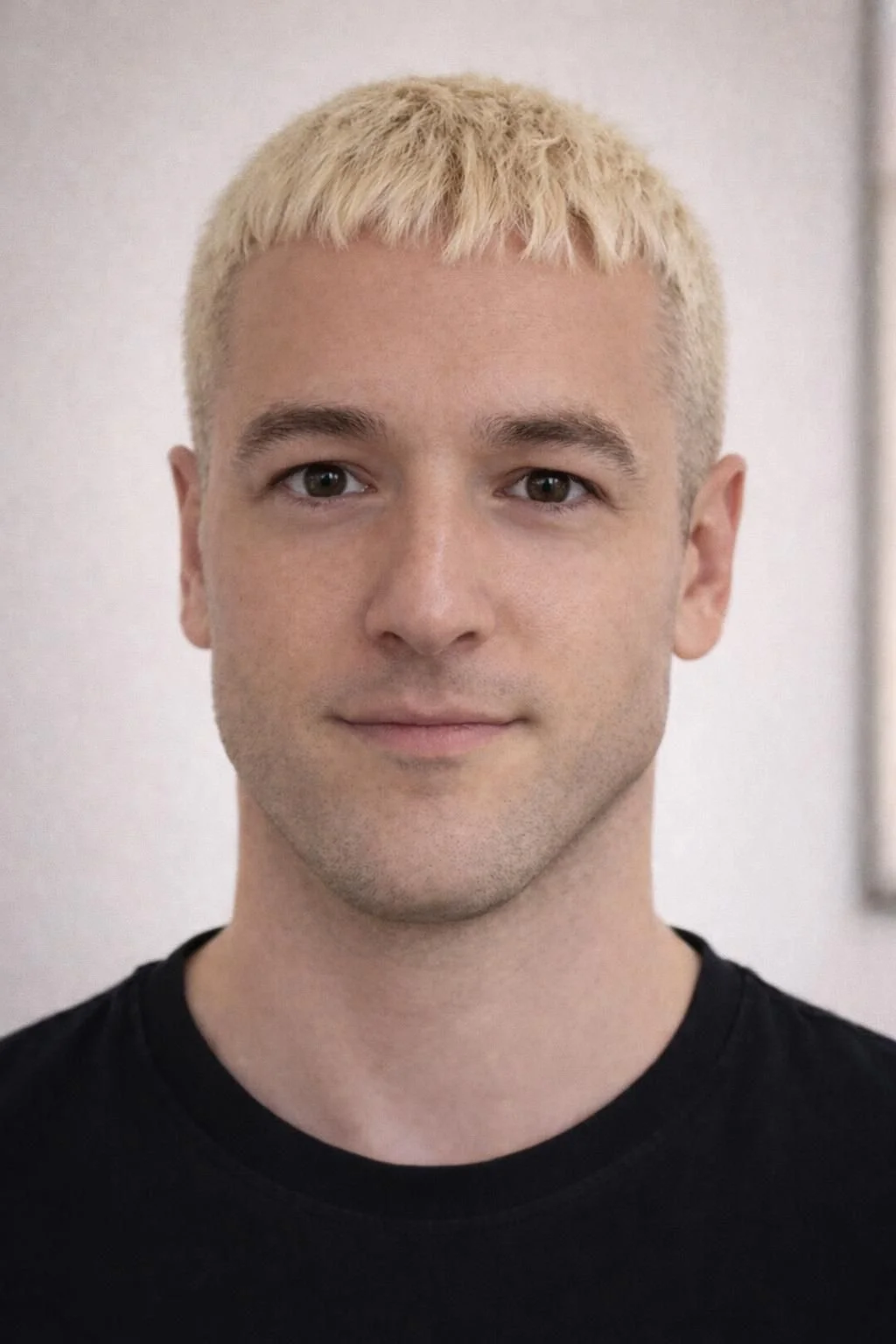 Retrato de un hombre joven con cabello rubio platinado, piel clara, ojos grandes y expresión neutral, usando una camiseta negra.