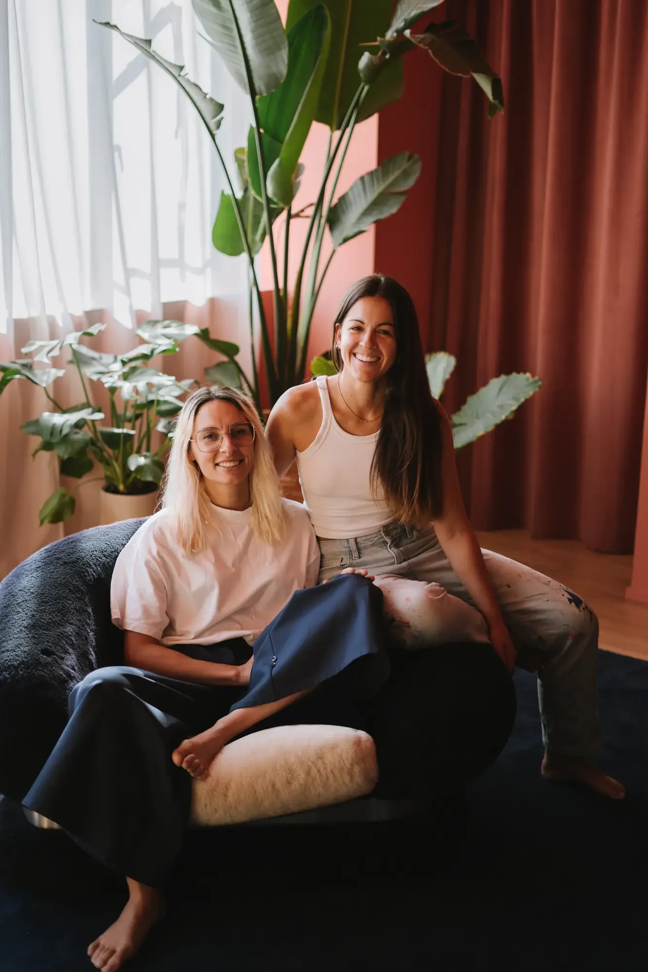 Dos mujeres sentadas en un sofá en una habitación con cortinas blancas y sotanás de cortina de color rosa y marrón, rodeadas de plantas verdes grandes, una de las mujeres lleva una blusa blanca y gafas, y la otra lleva una camiseta blanca y pantalones rotos. Ambas sonríen y parecen felices.