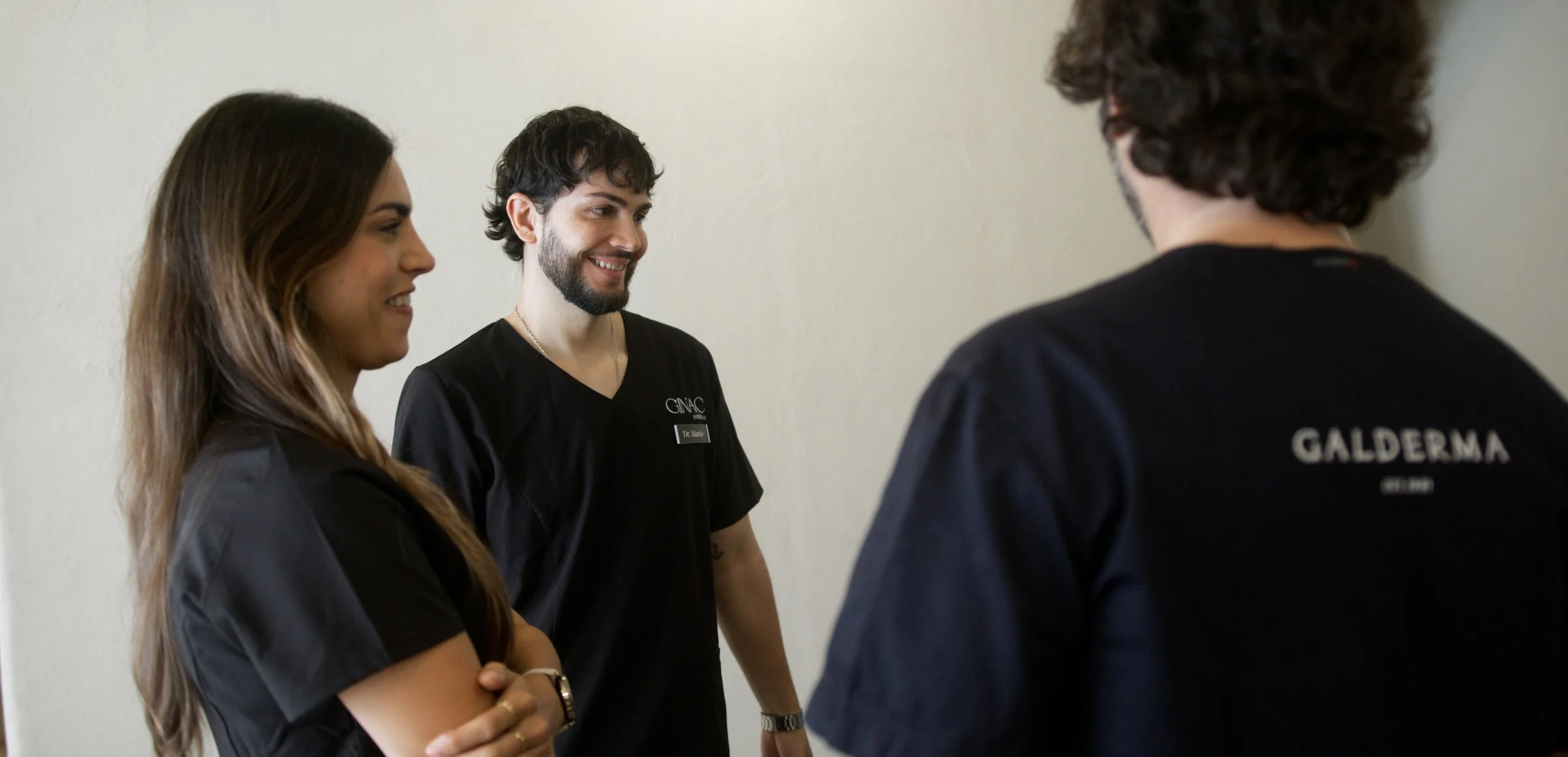 Tres profesionales de la salud conversando en una clínica, uno de ellos tiene una bata con el nombre 'GNAAC' y otro con 'GALDERMA' en la parte trasera.