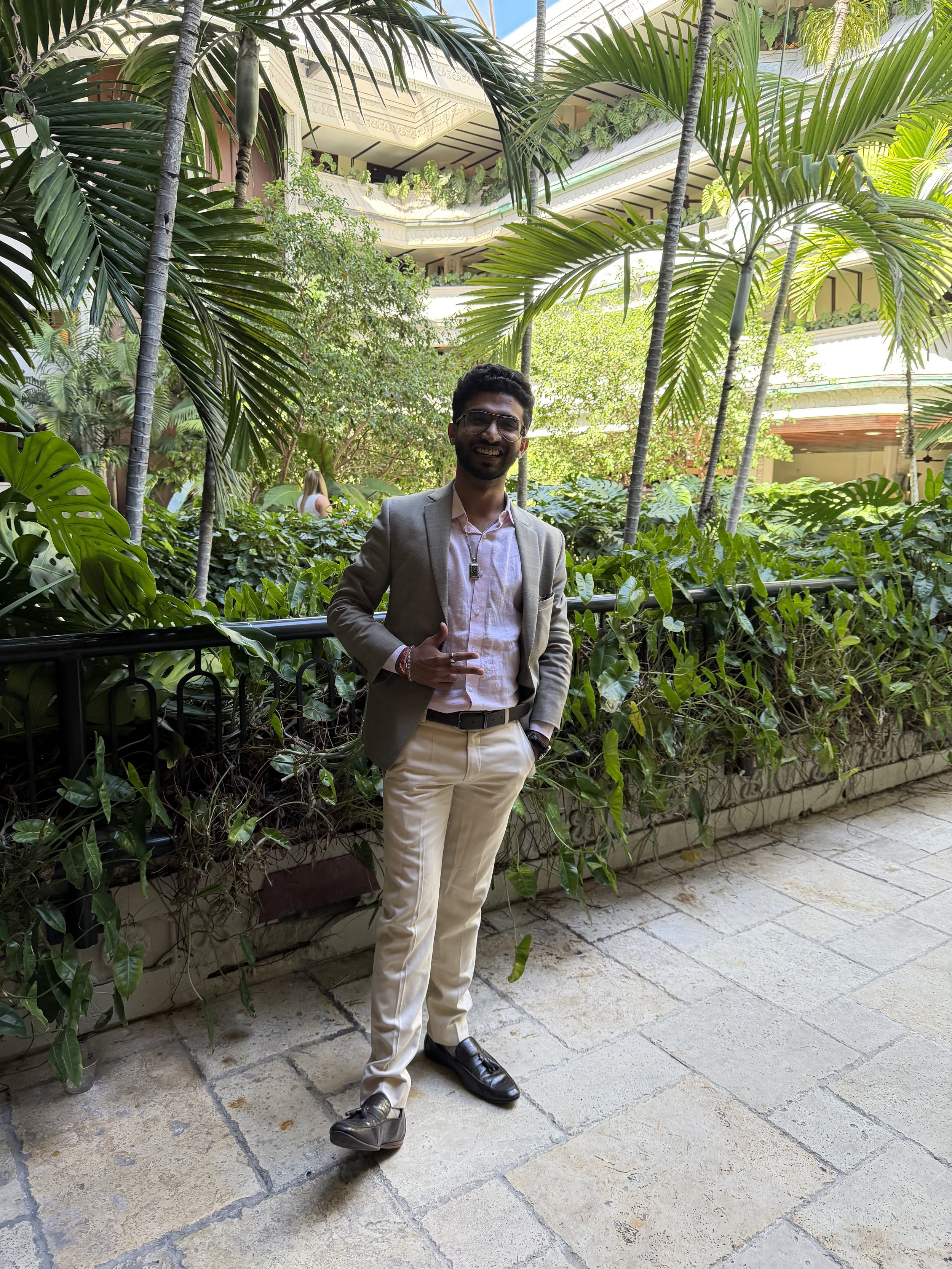 A smiling man in a beige suit and white shirt giving a thumbs-up, standing outdoors surrounded by lush green tropical plants, with multiple balconies and decorative elements in the background.