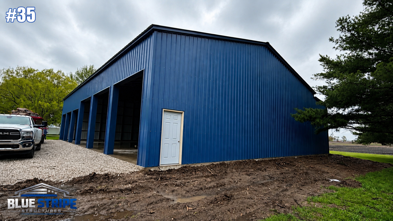 Large blue metal building with a white door, open bays, a gray gravel ramp, a pickup truck nearby, and trees in the background under cloudy skies.