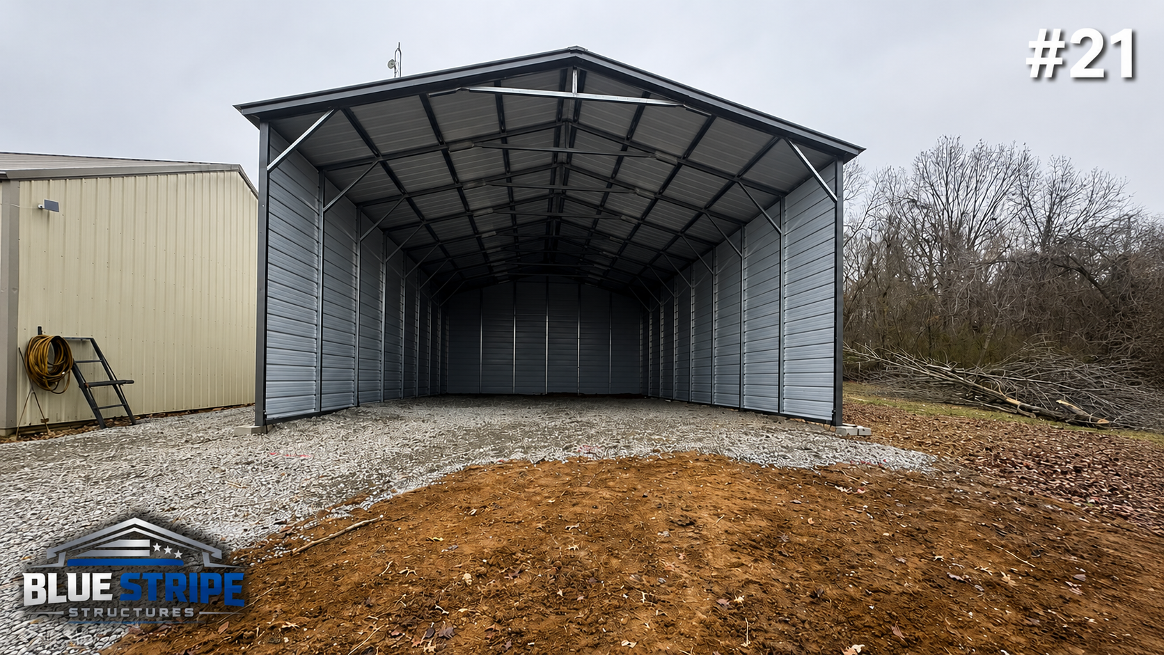 An empty metal storage shed with a curved roof, open front, and gray siding, situated outdoors on gravel ground, with a cream-colored building on its left side and leafless trees in the background.