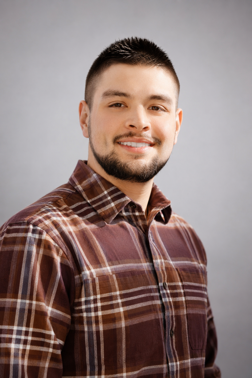 Portrait of a young man with short dark hair and a beard, smiling, wearing a brown plaid shirt, standing against a gray background.