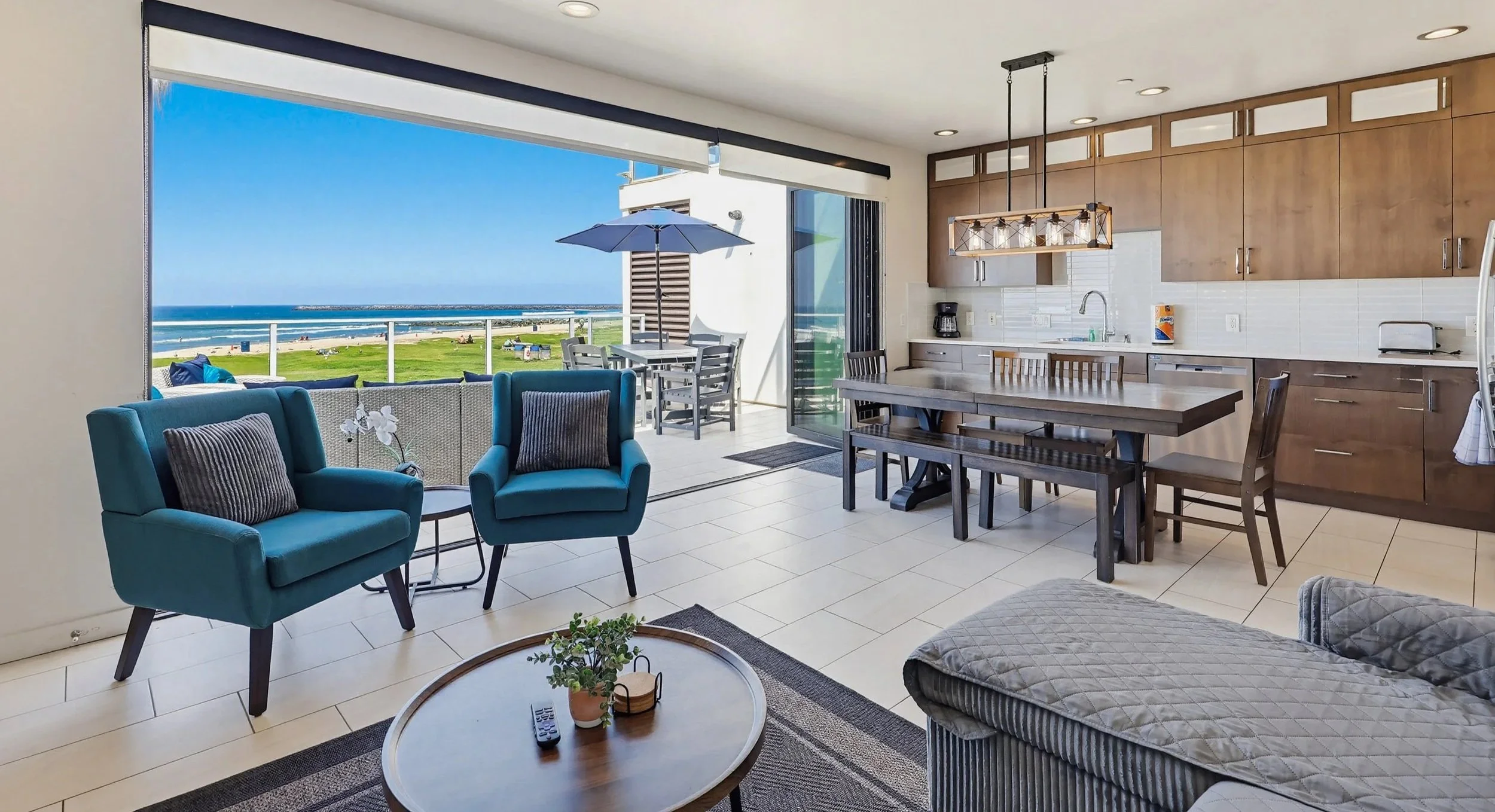 Living room with open view to outdoor balcony overlooking the beach, with seating and dining areas, wooden cabinets in the kitchen, and a view of blue sky and ocean.