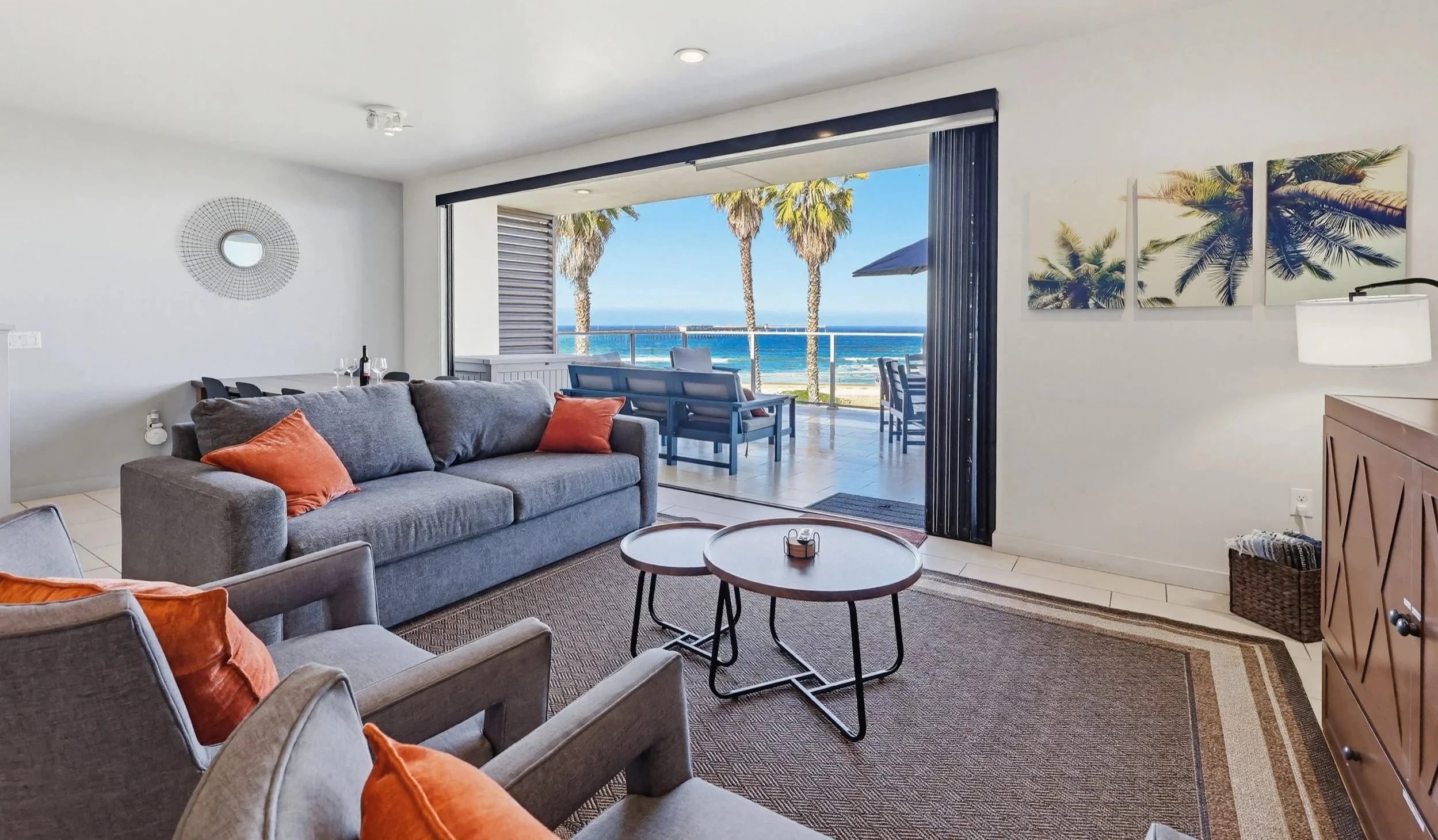 Living room with a view of the ocean, featuring a gray sofa with orange pillows, a coffee table, and artwork of palm trees on the wall. The room opens to an outdoor balcony with chairs and a table, overlooking palm trees and the ocean.