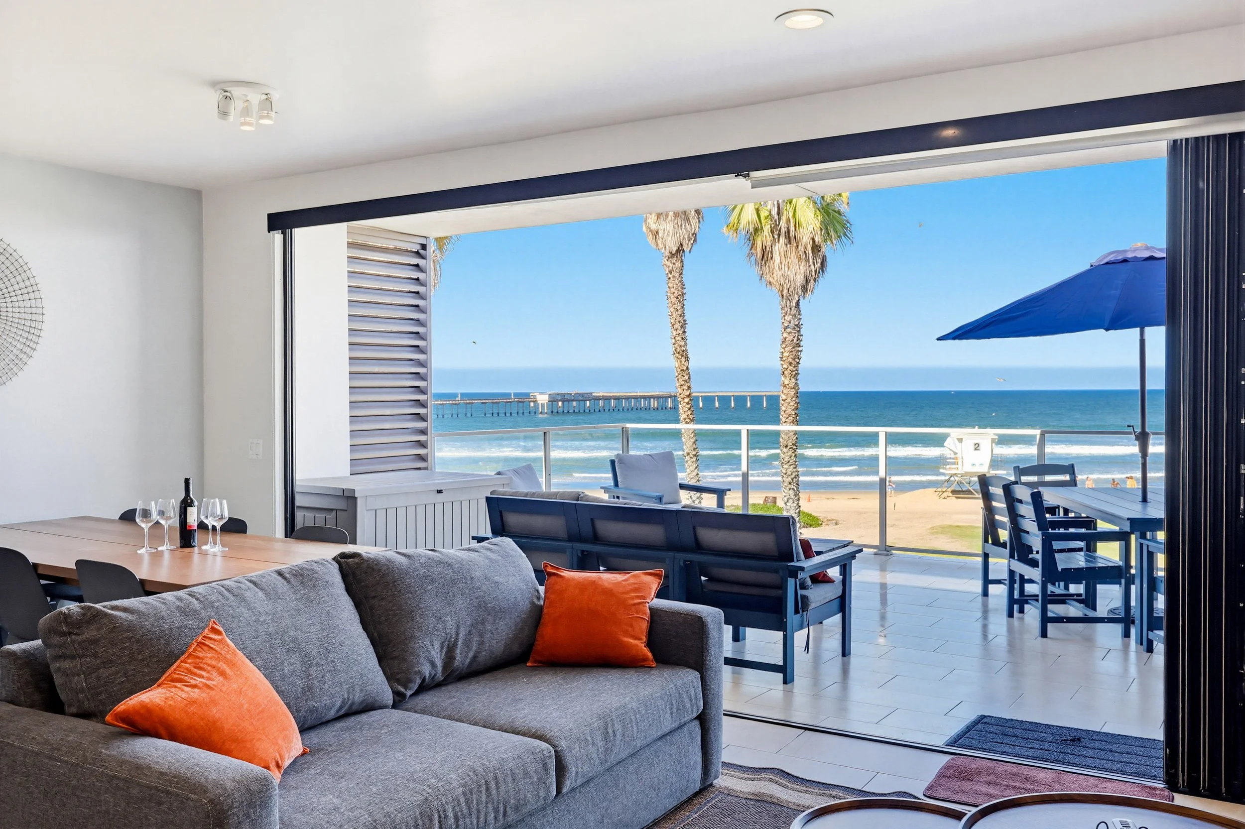 Living room with a large open window showing a beach scene with sand, ocean, palm trees, outdoor furniture, and a pier in the background.