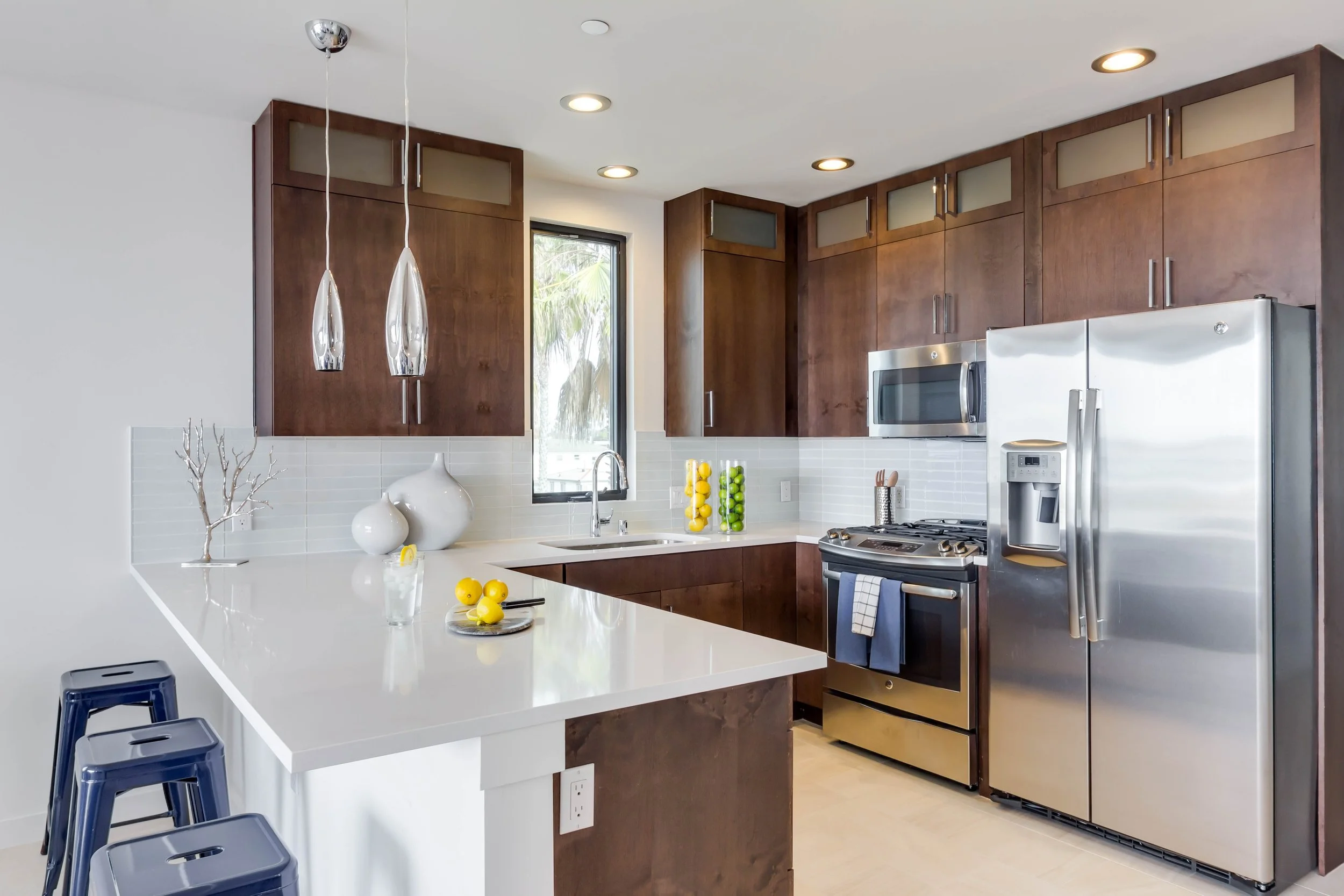 Modern kitchen with dark wood cabinets, stainless steel appliances, white countertops, and white subway tile backsplash. Decorative vases and lemons are on the counter, with pendant lights hanging above and a window behind the sink.