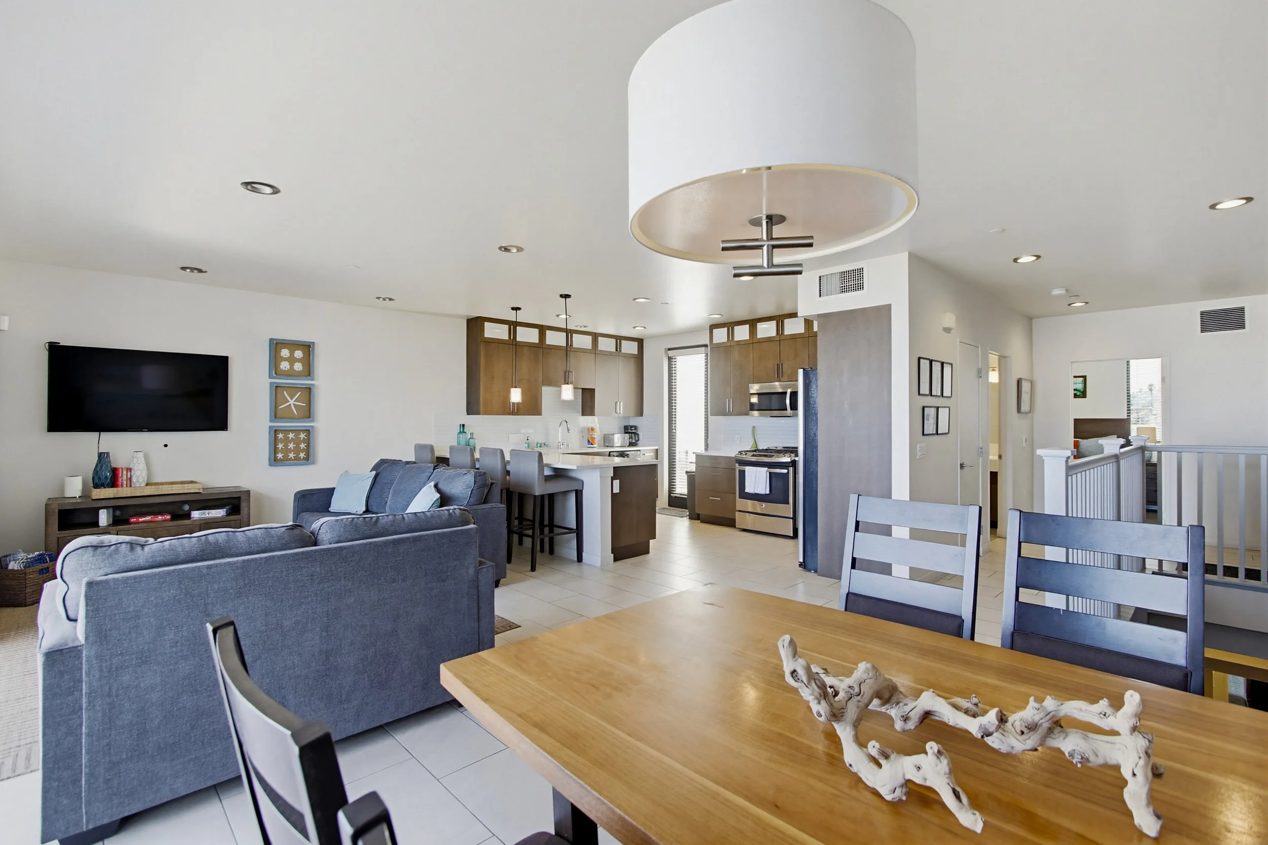 Open-plan living room and kitchen area with grey sofas, wooden dining table with a branch centerpiece, and modern kitchen cabinets.