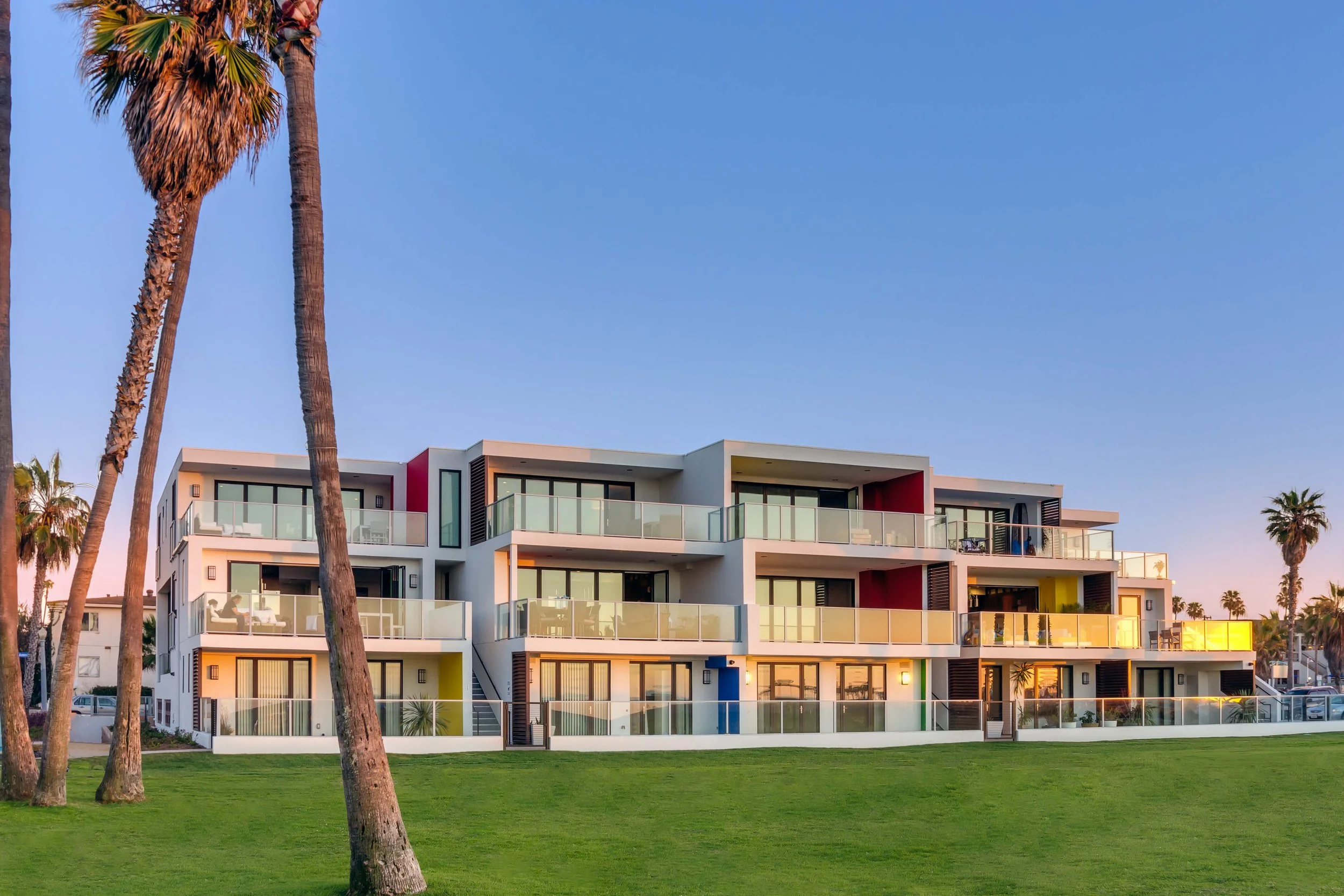 A modern, multi-story vacation rentals building with large glass balconies, set against a clear blue sky and surrounded by tall palm trees and a lush green lawn.