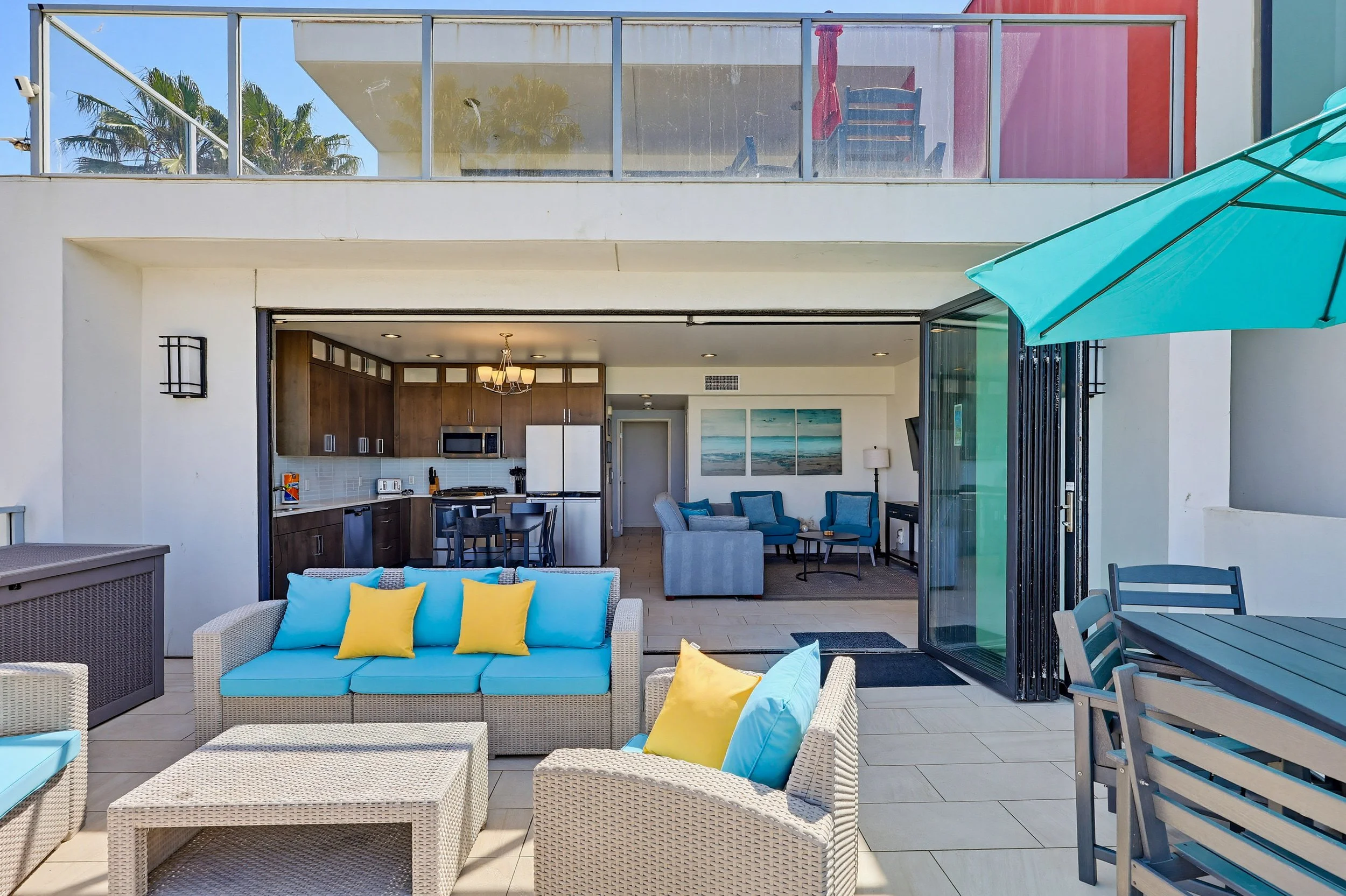 Outdoor patio with wicker furniture, blue and yellow cushions, dining table, blue patio umbrella, and partially open sliding glass door revealing a living room with modern decor and kitchen area.