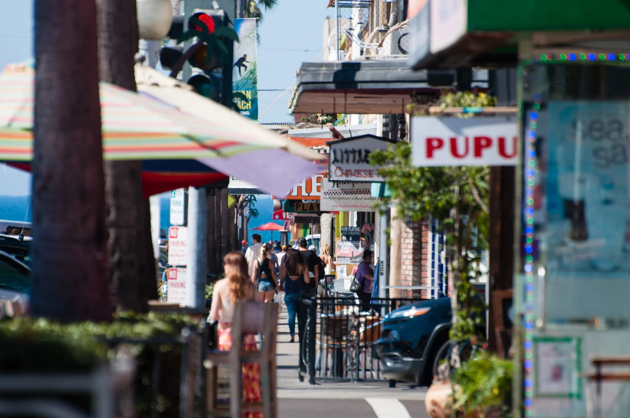 A busy street with pedestrians walking along the sidewalk, lined with various shops and storefronts. Signage for businesses is visible, including a sign that says 'Pupu.' A colorful striped umbrella is partially visible in the foreground, and there are trees and utility poles along the street.