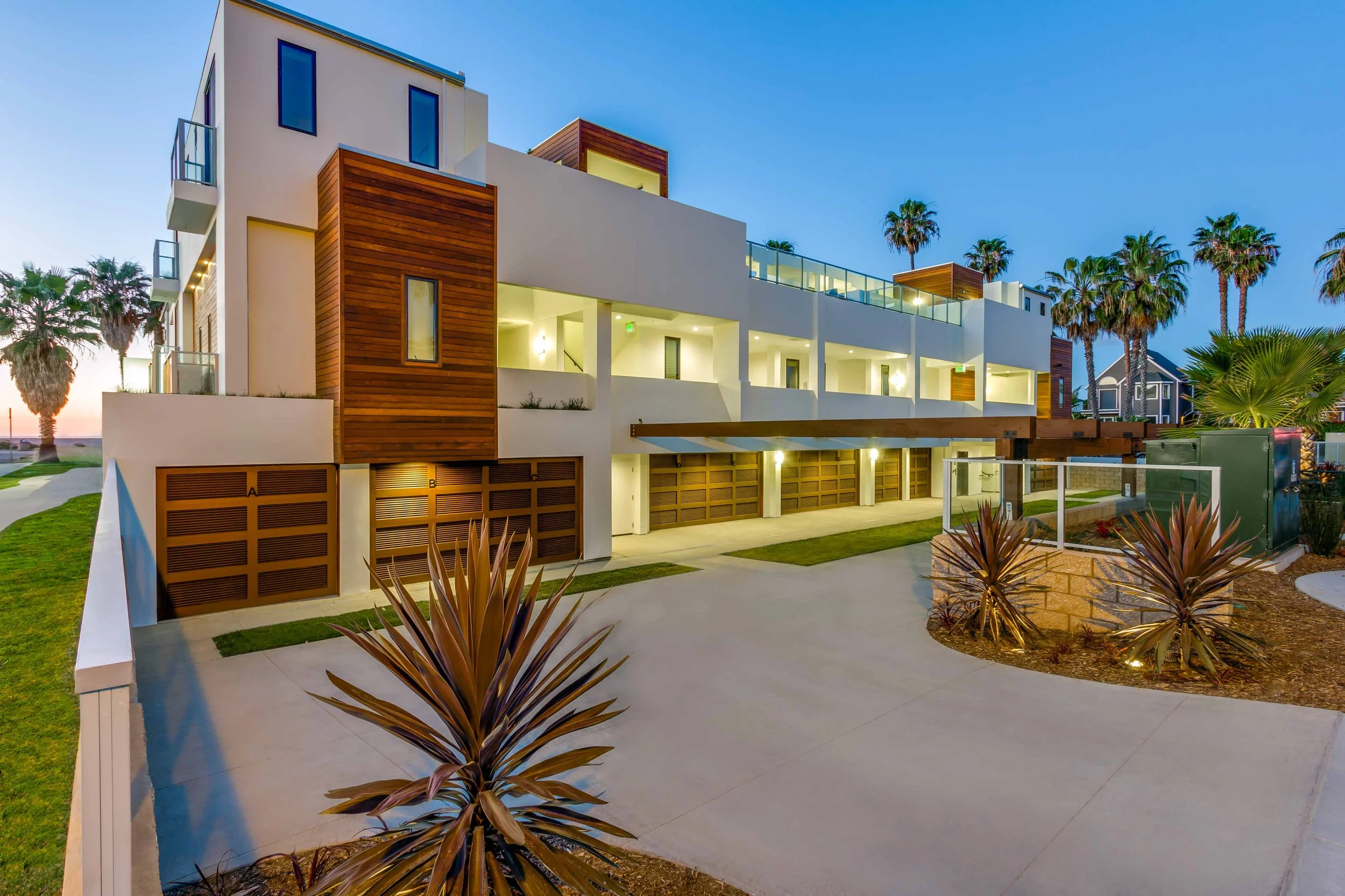 Modern multi-story vacation rentals building with white walls, wooden accents, glass balconies, and palm trees, illuminated by outdoor lighting during dusk.