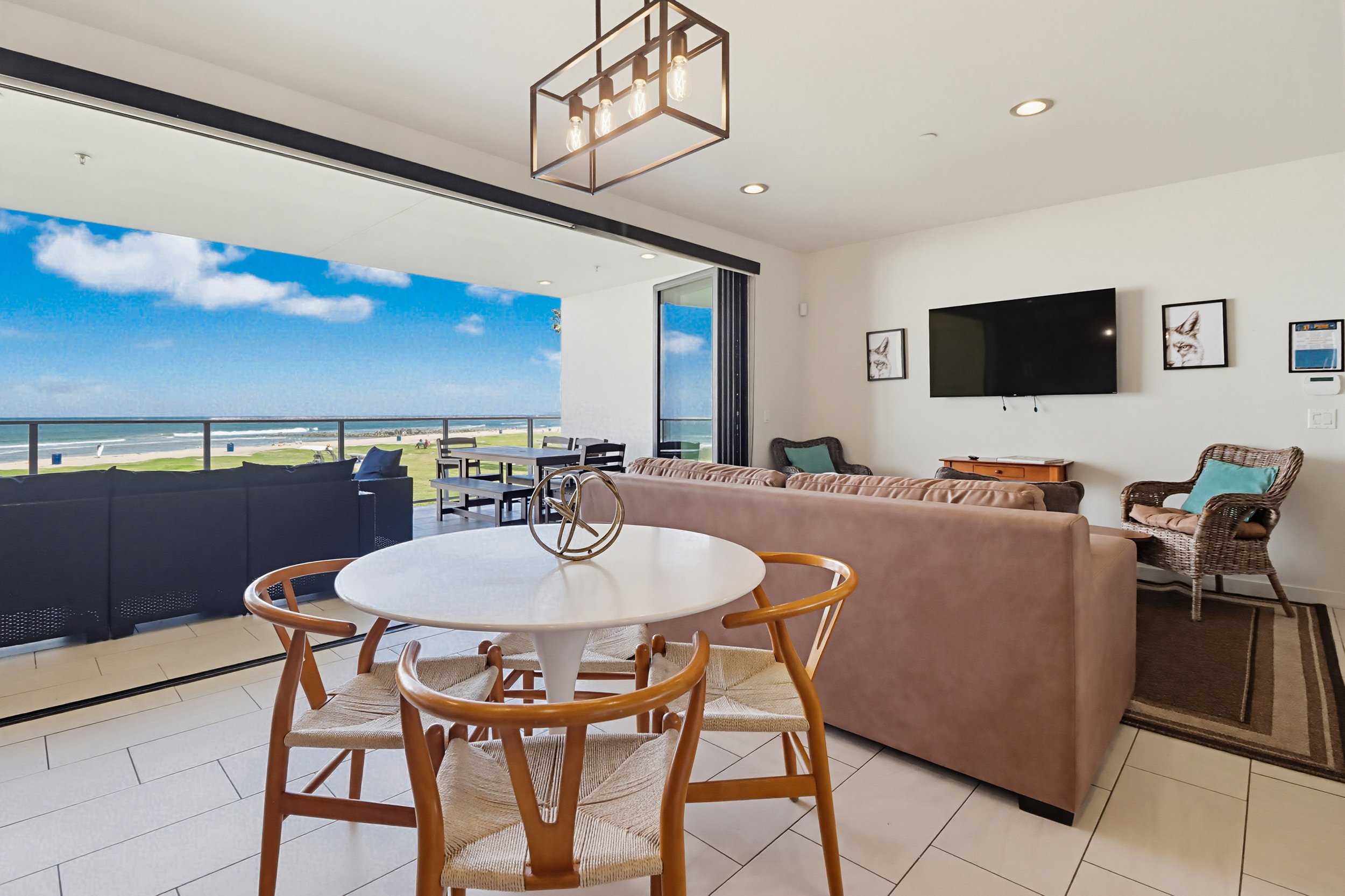 A living room with a view of the beach and ocean through a large sliding door, featuring furniture including a round dining table with wooden chairs, sofas, and chairs, a wall-mounted flat-screen TV, framed artwork, and modern lighting fixtures.