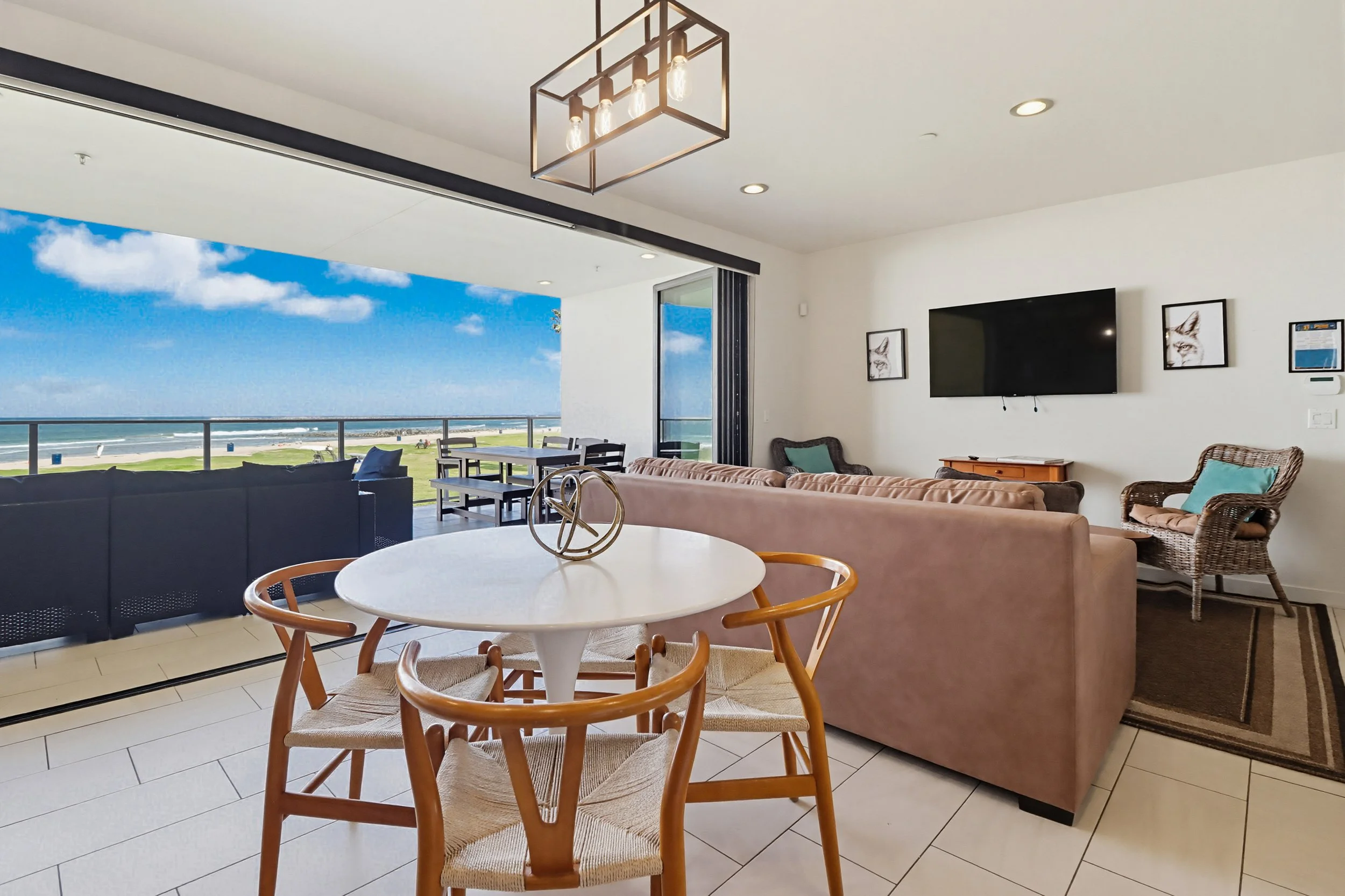 A living room with a view of the beach and ocean through a large sliding door, featuring furniture including a round dining table with wooden chairs, sofas, and chairs, a wall-mounted flat-screen TV, framed artwork, and modern lighting fixtures.
