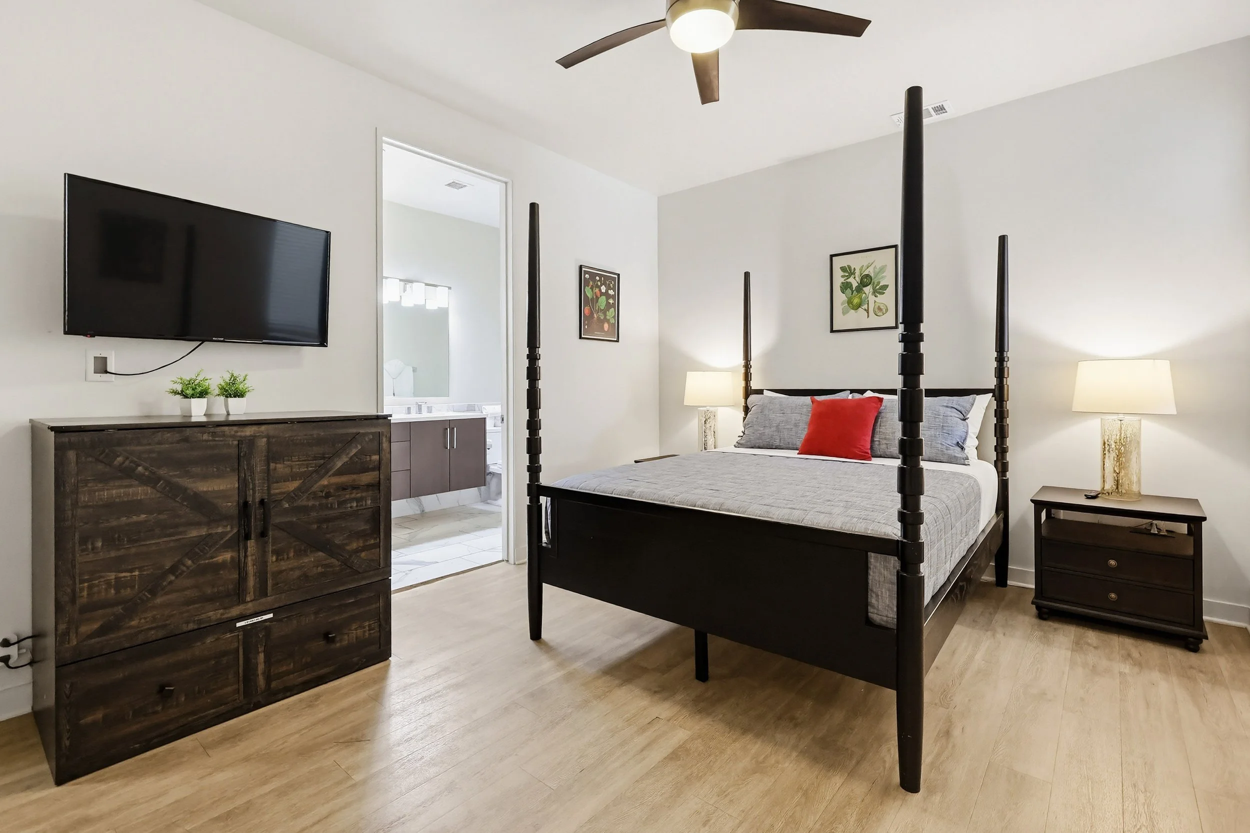 A bedroom with a black four-poster bed with gray bedding and a red pillow, two nightstands with lamps, a wooden dresser, a wall-mounted flat-screen television, and framed botanical prints, with an open door leading to a bathroom.