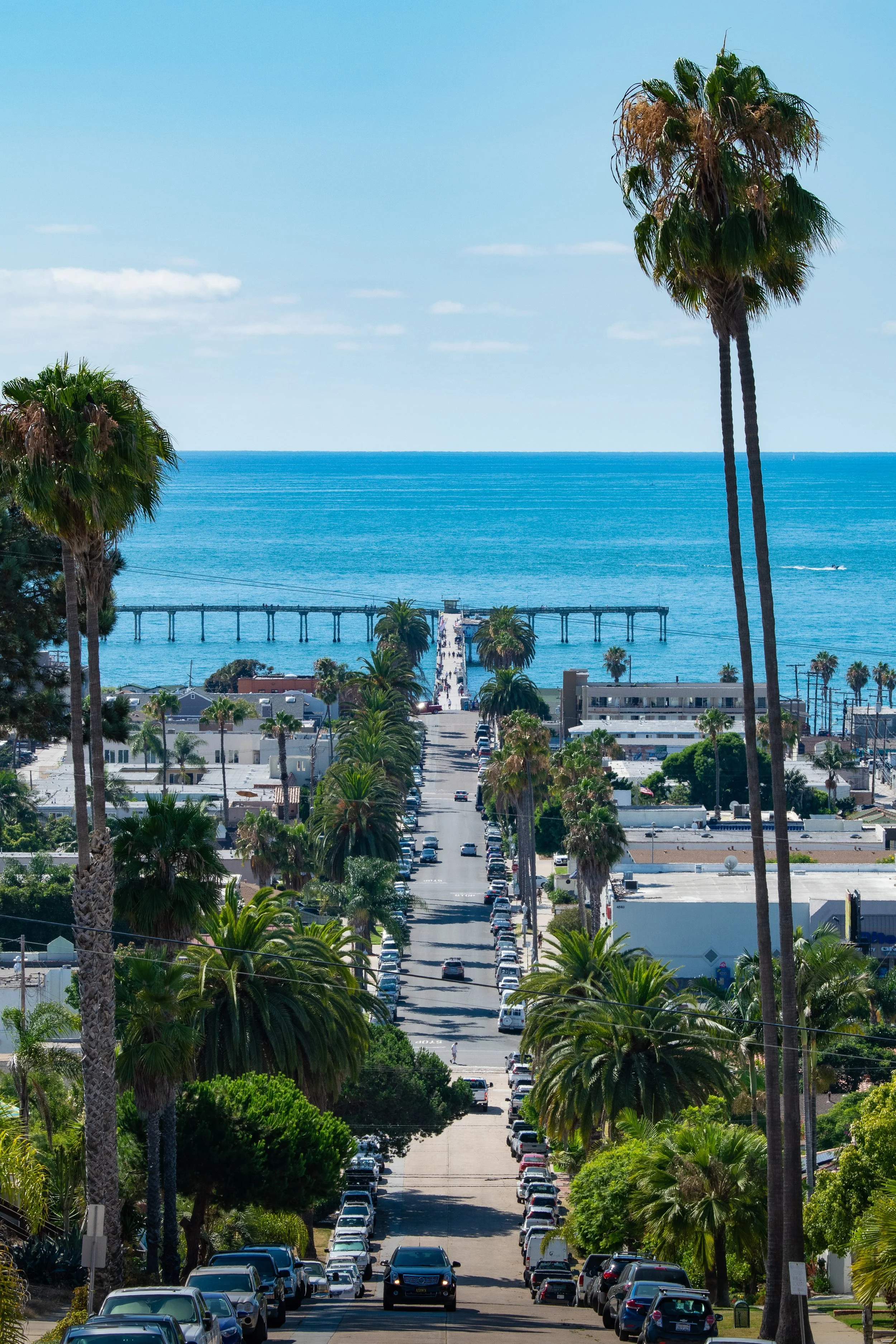 A view of a seaside street in Ocean Beach, CA with palm trees, leading to a pier and the ocean under a partly cloudy sky.
