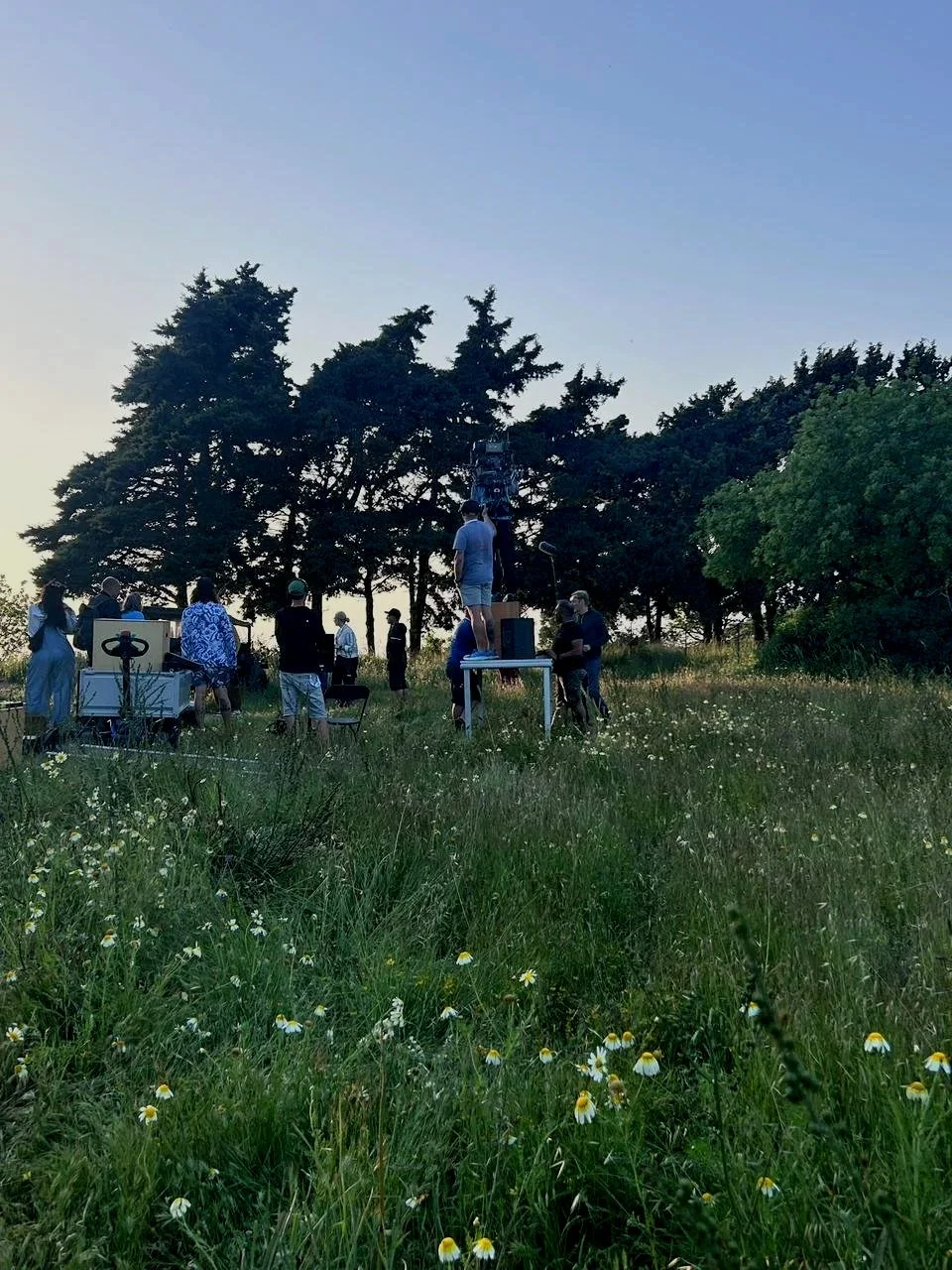 Group of people outdoors in a grassy field with wildflowers, some gathered around equipment and a tall camera mounted on a stand, during the evening or late afternoon.