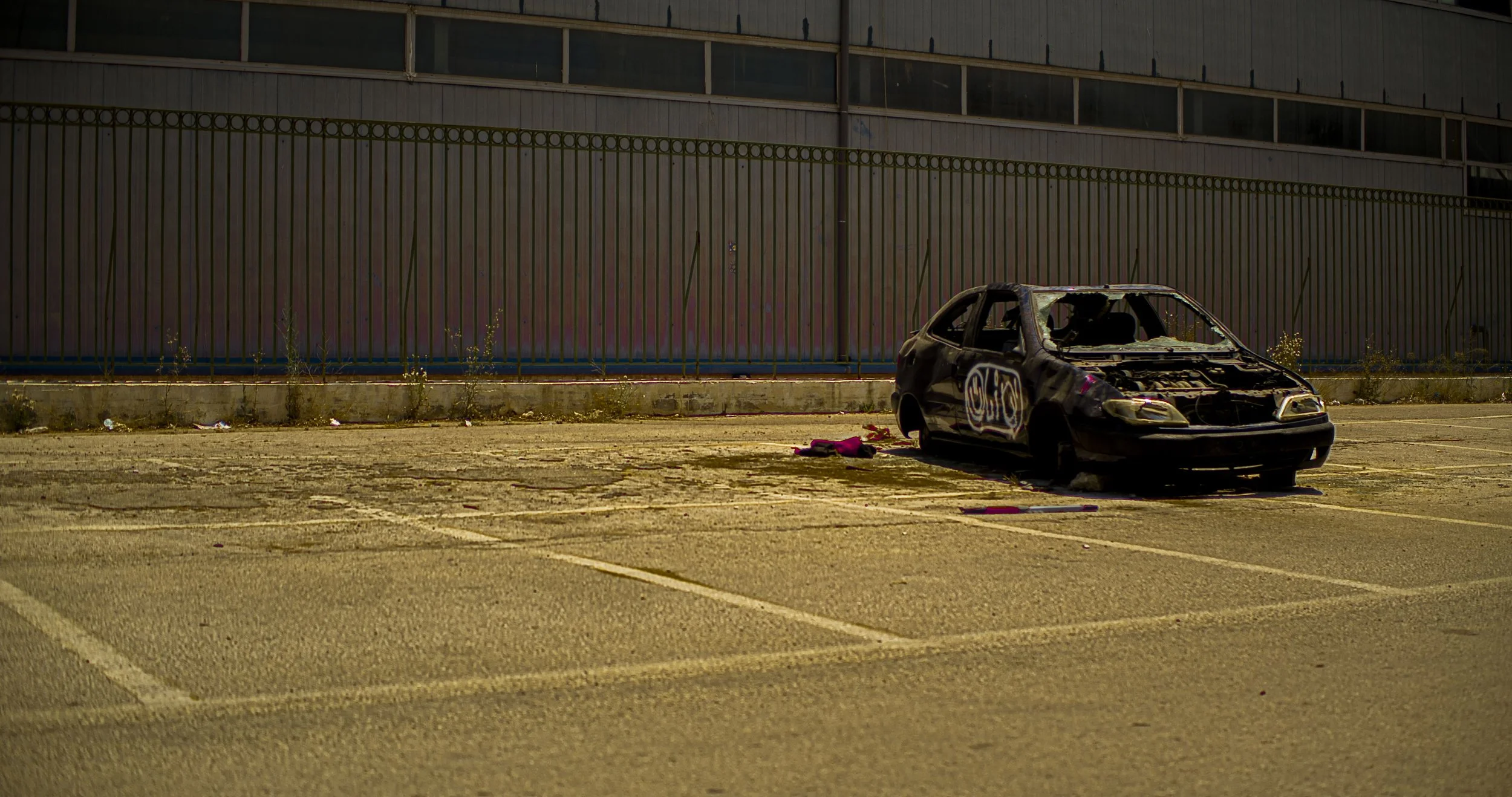 Burned-out black car in an empty parking lot with graffiti on the side, behind a tall metal fence.
