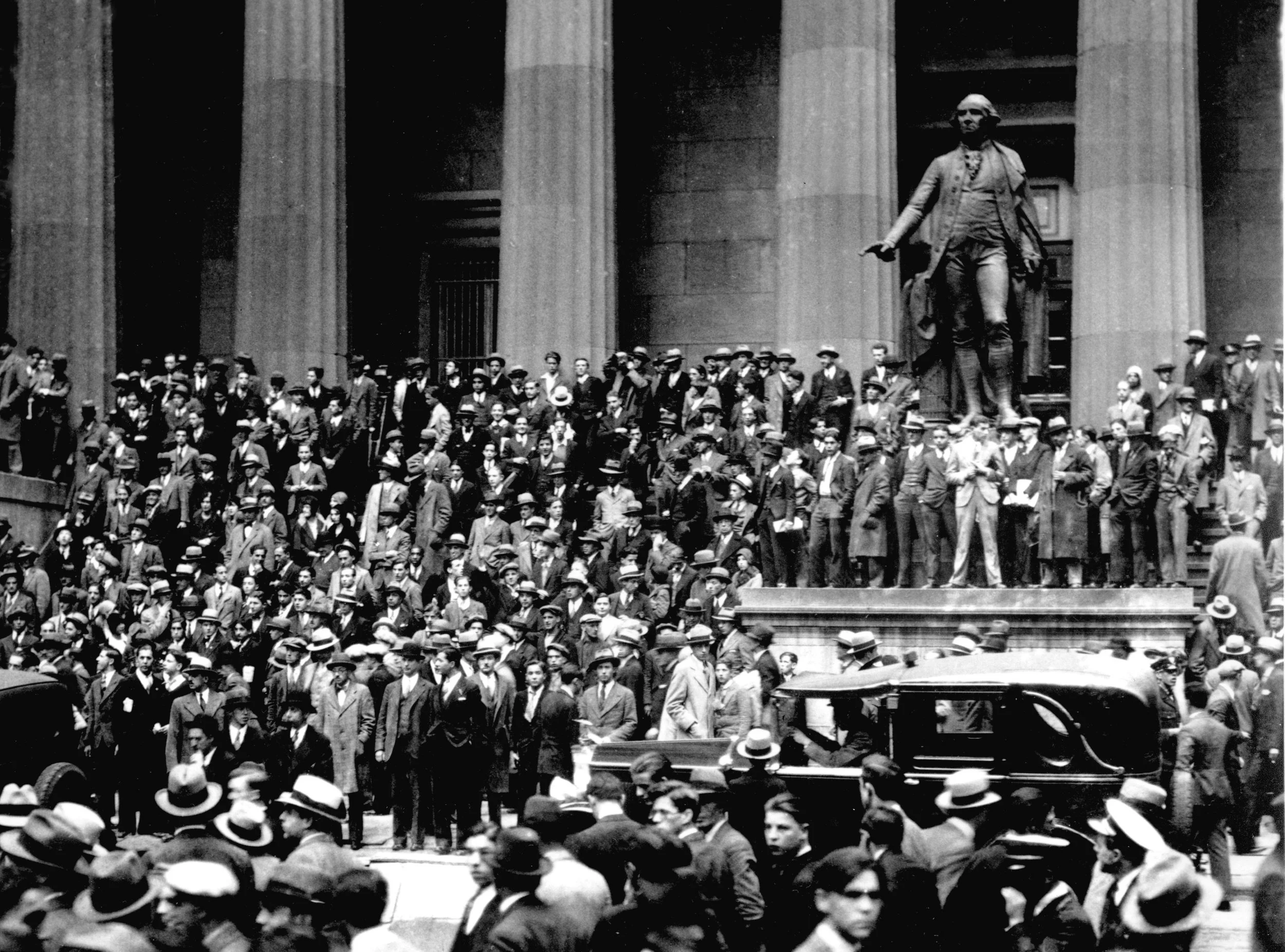 A black-and-white historical photograph of a large crowd of men gathered outside a building with tall columns. The men are dressed in early 20th-century attire, wearing suits, ties, and hats. A prominent statue of a man stands in the middle of the scene.