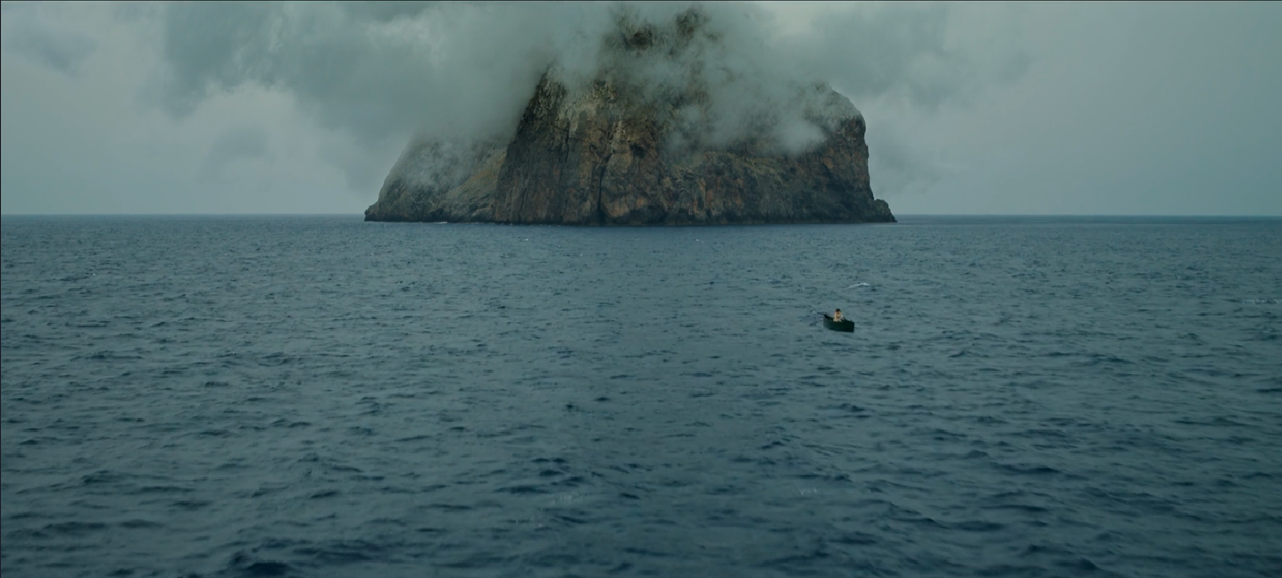 A small boat with one person on it floating on the ocean, with a large rocky island shrouded in clouds in the background.