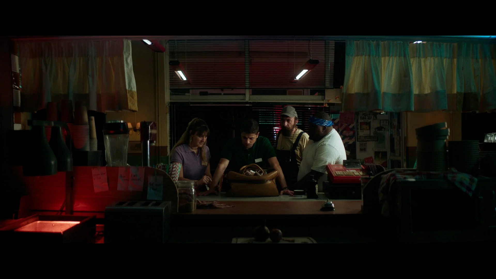 Four people working behind a counter at a small eatery, with a woman and a man at the front of the counter, and two other men in the background. The counter is filled with cups, pitchers, and a cash register. The scene is dimly lit with colorful curt
