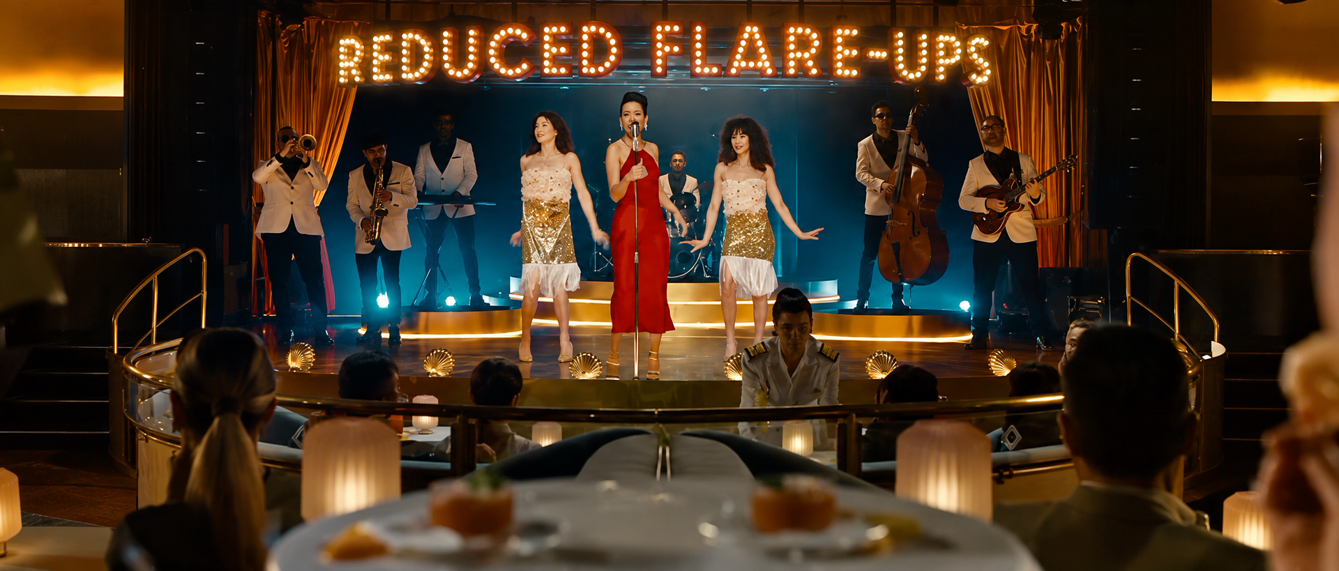 A musical performance on stage with women in elegant dresses and band members in suits, with a sign reading 'Reduced Flare-Ups' above.
