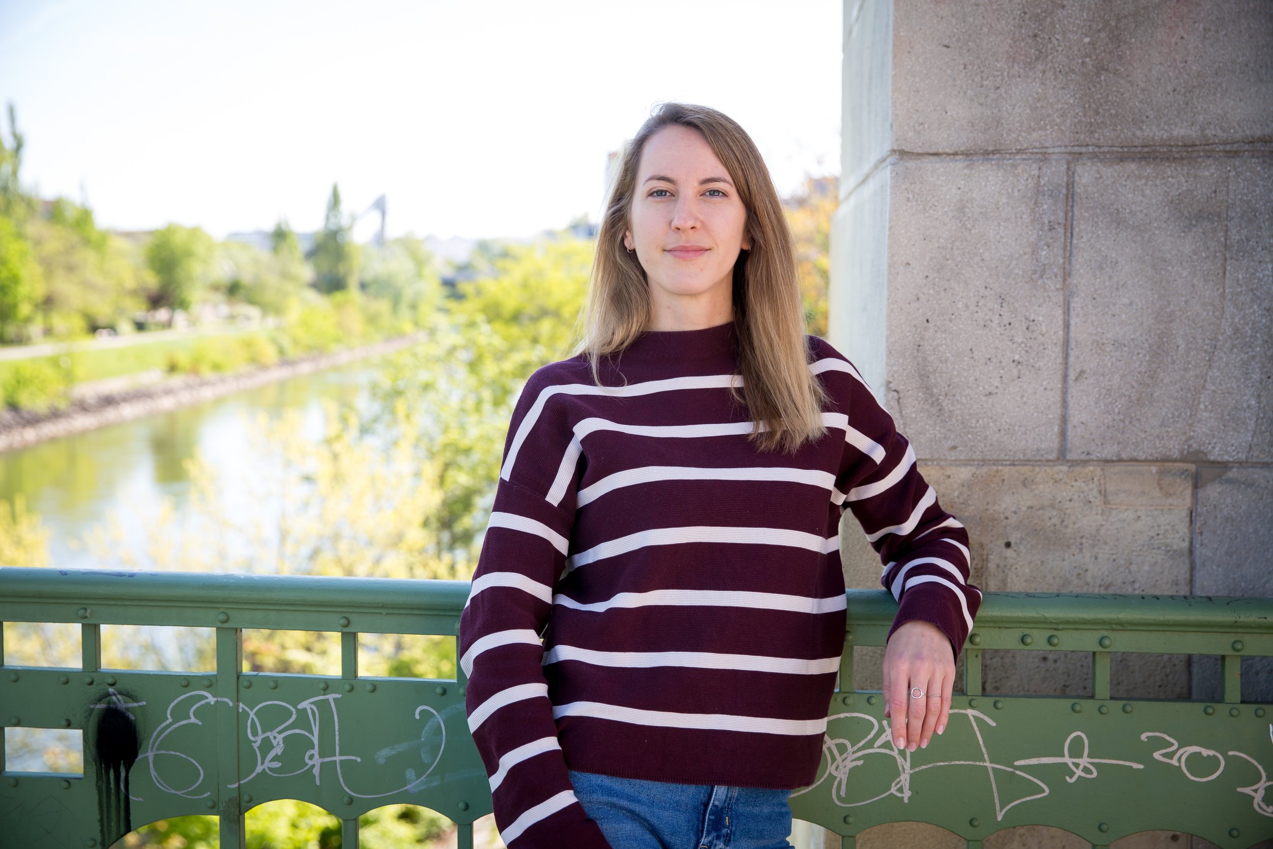 Personzentrierte Psychotherapeutin steht auf einer Brücke mit Graffiti in Wien, trägt ein lila-weiß gestreiftes Oberteil und blaue Jeans, im Hintergrund sind Bäume und Wasser sichtbar.