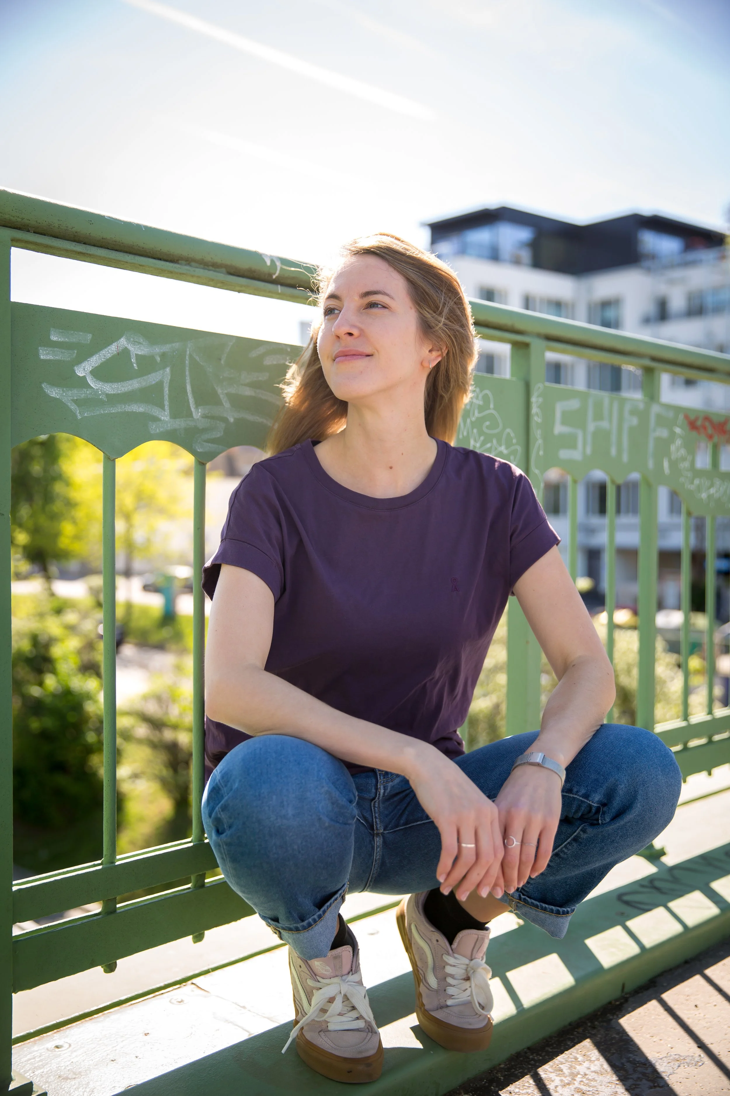 Personzentrierte Psychotherapeutin sitzt entspannt auf einer grünen Parkbank, trägt eine dunkle T-Shirt, blaue Jeans, Turnschuhe und schaut zur Seite bei Sonnenlicht. Im Hintergrund sind Wohnhäuser in Wien, Bäume und ein blauer Himmel sichtbar.