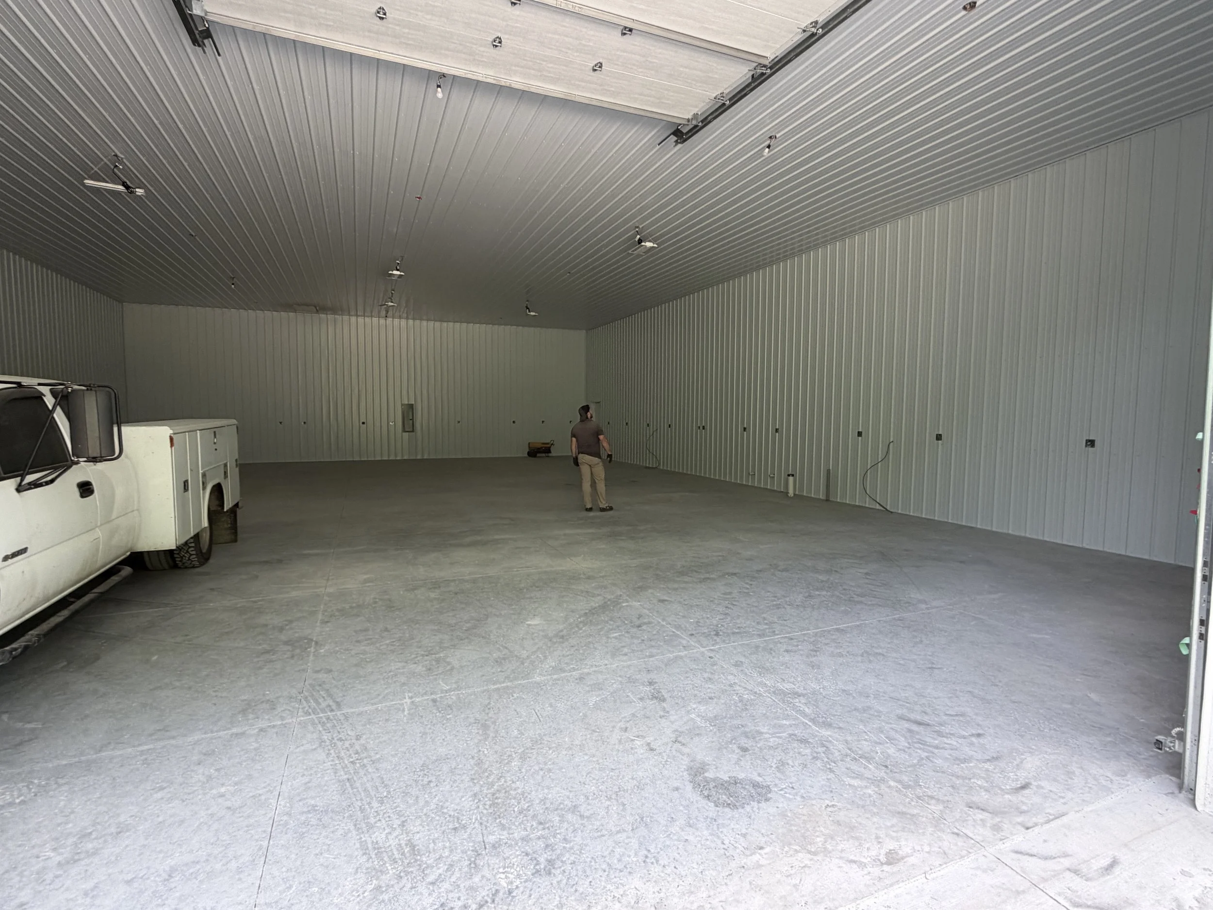 Interior of an unfinished large metal building with a high ceiling, concrete floor, and walls made of metal panels. There are several light fixtures hanging from the ceiling and a person working near the back wall, along with a white utility truck parked inside.