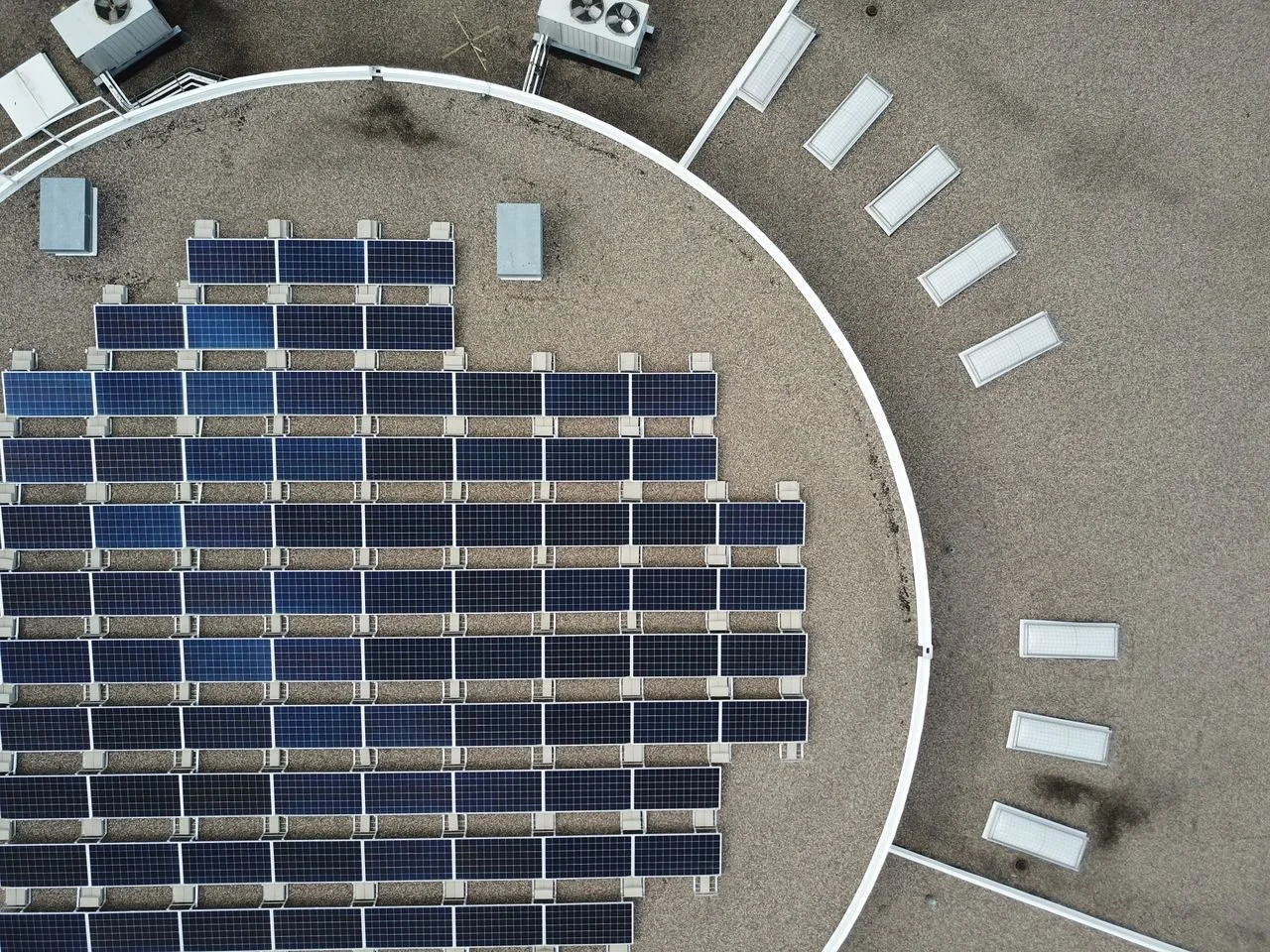 Aerial view of a solar panel installation on a flat rooftop, with solar panels arranged in rows and white rectangular skylights on the surrounding gravel surface.