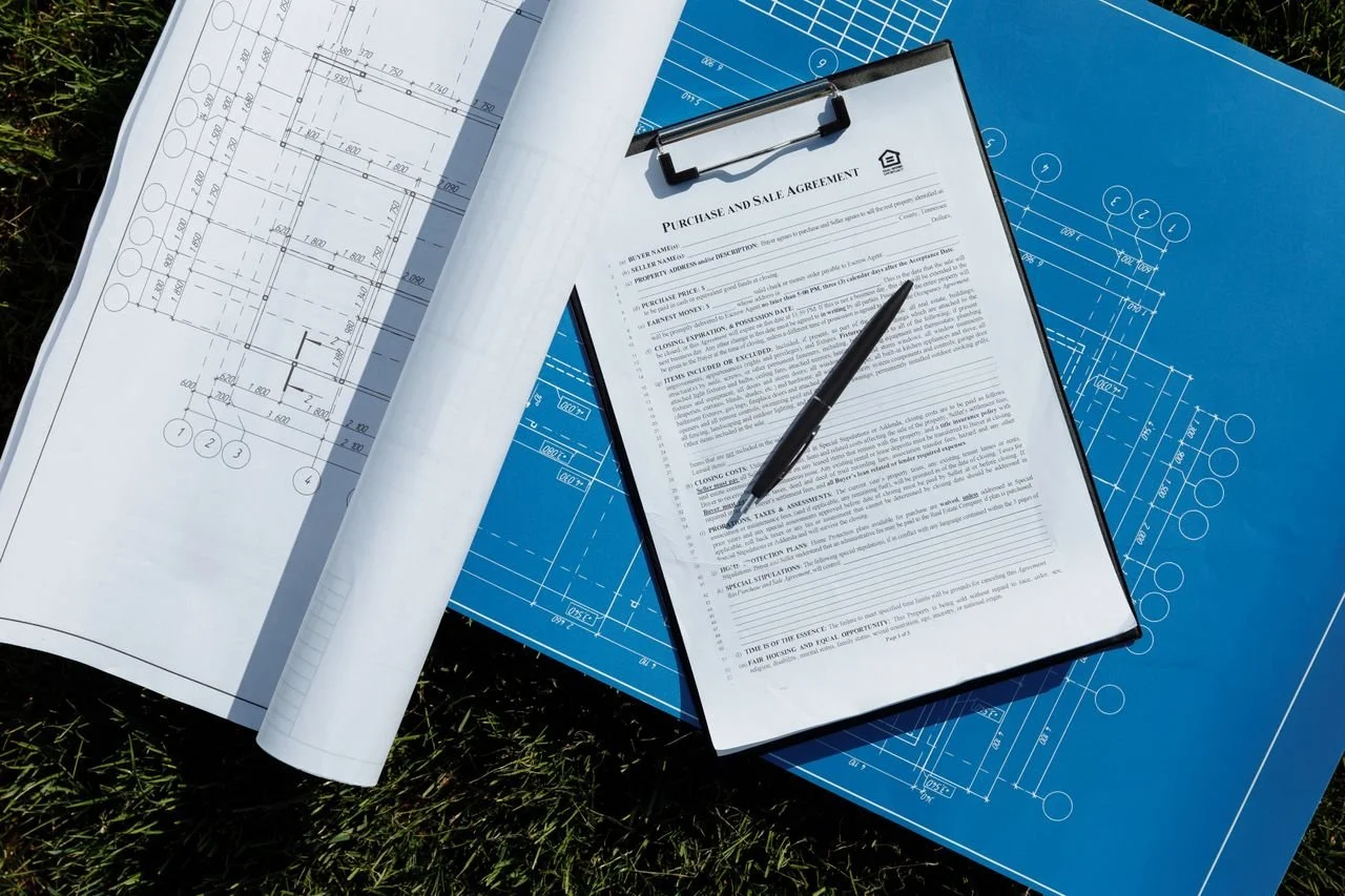 Blueprint drawings on blueprints and white paper, a clipboard with a purchase and sale agreement, and a black pen on top of the papers, placed outdoors on grass.