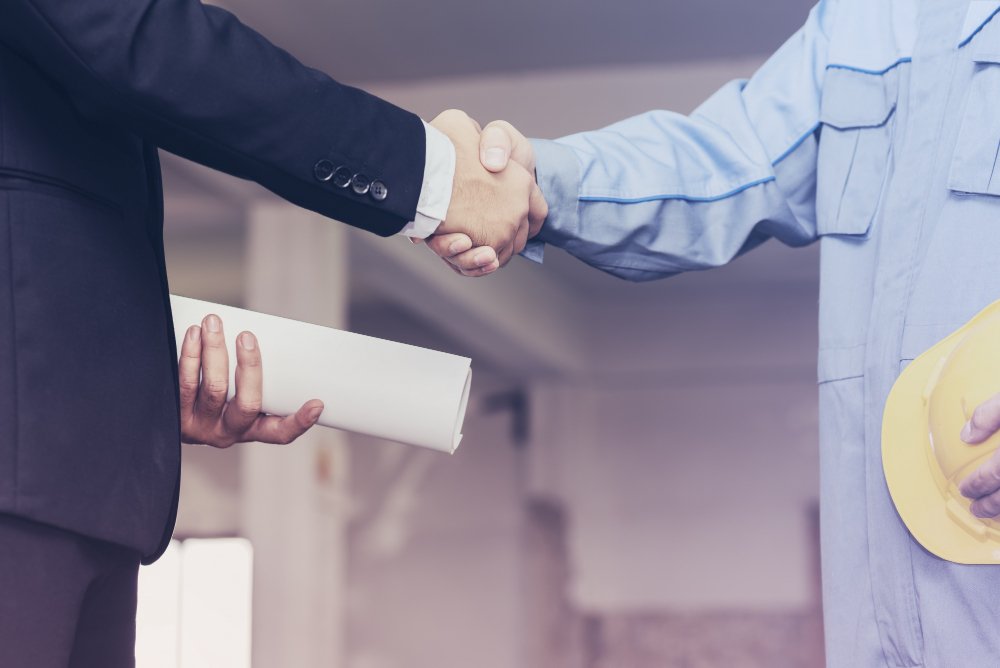 Two people shaking hands, one in a suit holding a rolled document, the other in a work uniform holding a yellow hard hat.